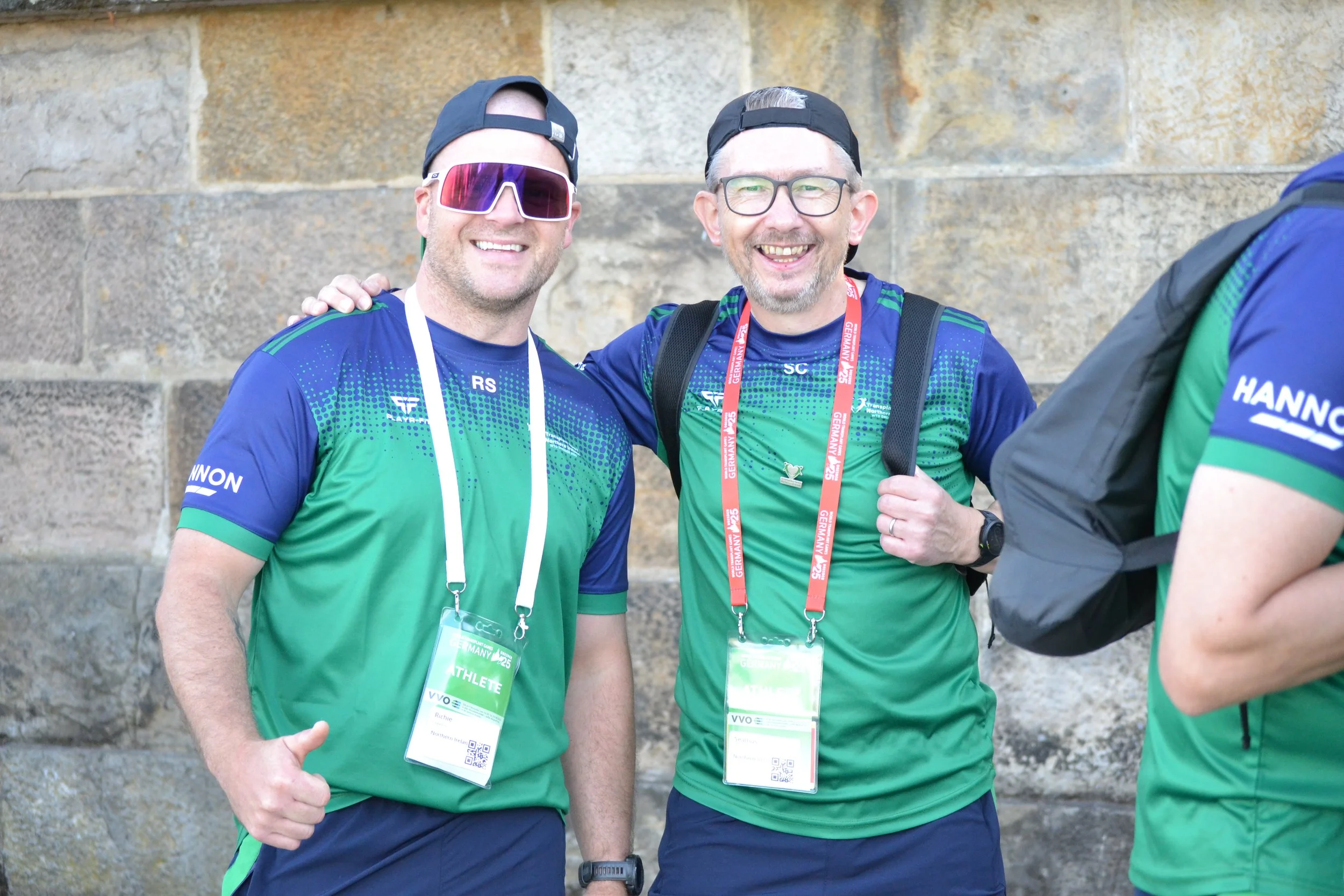 Two smiling men wearing athletic gear and medals around their necks, posing outdoors against a stone wall. One man is wearing sunglasses and a black cap, while the other has glasses and a black cap worn backwards.