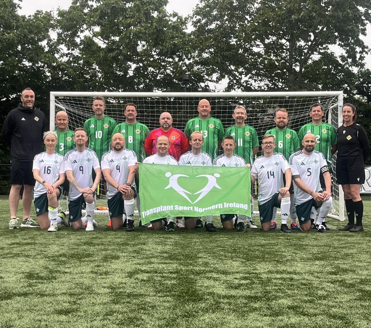Group of people, some wearing green and white soccer uniforms, posing in front of a soccer net, holding a green banner that reads 'Transplant Sport Northern Ireland', on a grassy field with trees in the background.