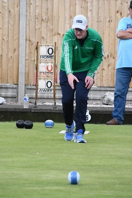 A person in a green jacket and white cap playing lawn bowls on a grass field, with other players and a scoreboard in the background.