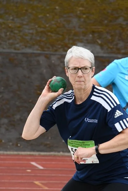 Older woman with short gray hair and glasses throwing a green shot put during a sporting event at an outdoor track.