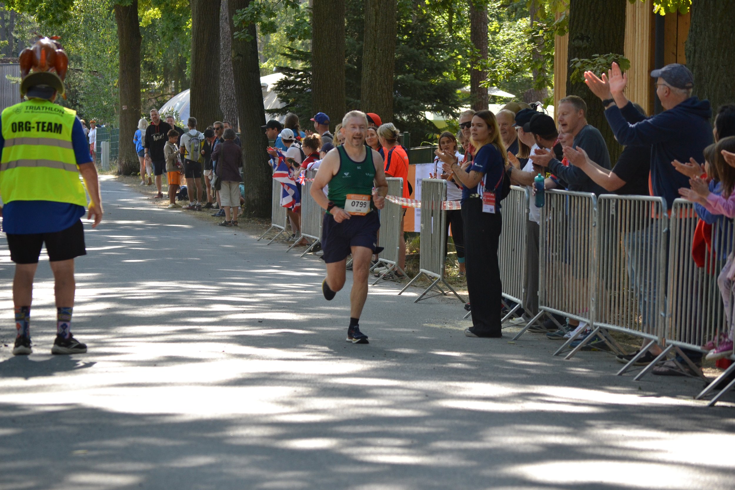 A marathon runner crosses the finish line while spectators cheer from behind a metal barricade in a wooded area.