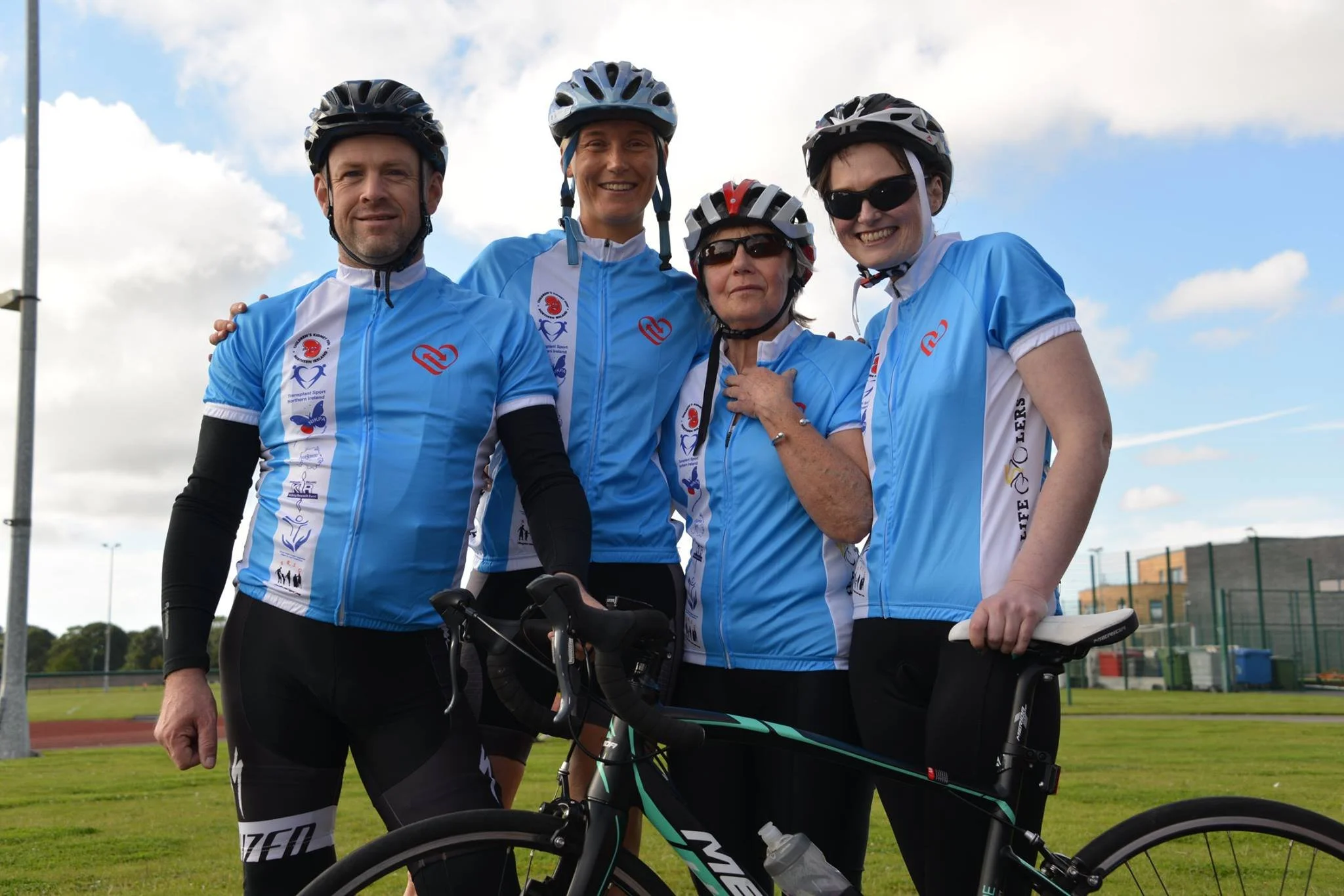 Group of five cyclists in blue jerseys and helmets smiling outdoors on a cloudy day, with a sports field and stadium in the background.