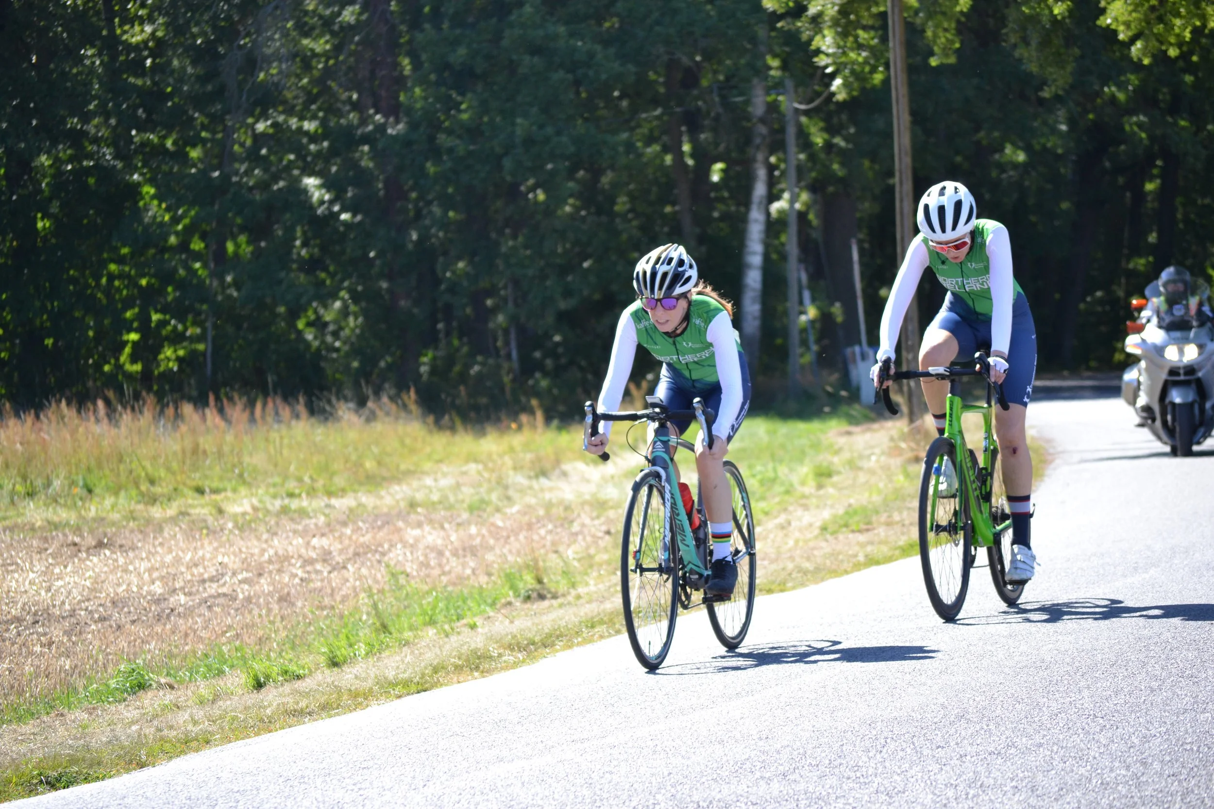 Two cyclists wearing green jerseys and helmets riding on a paved trail through a park or wooded area on a sunny day.