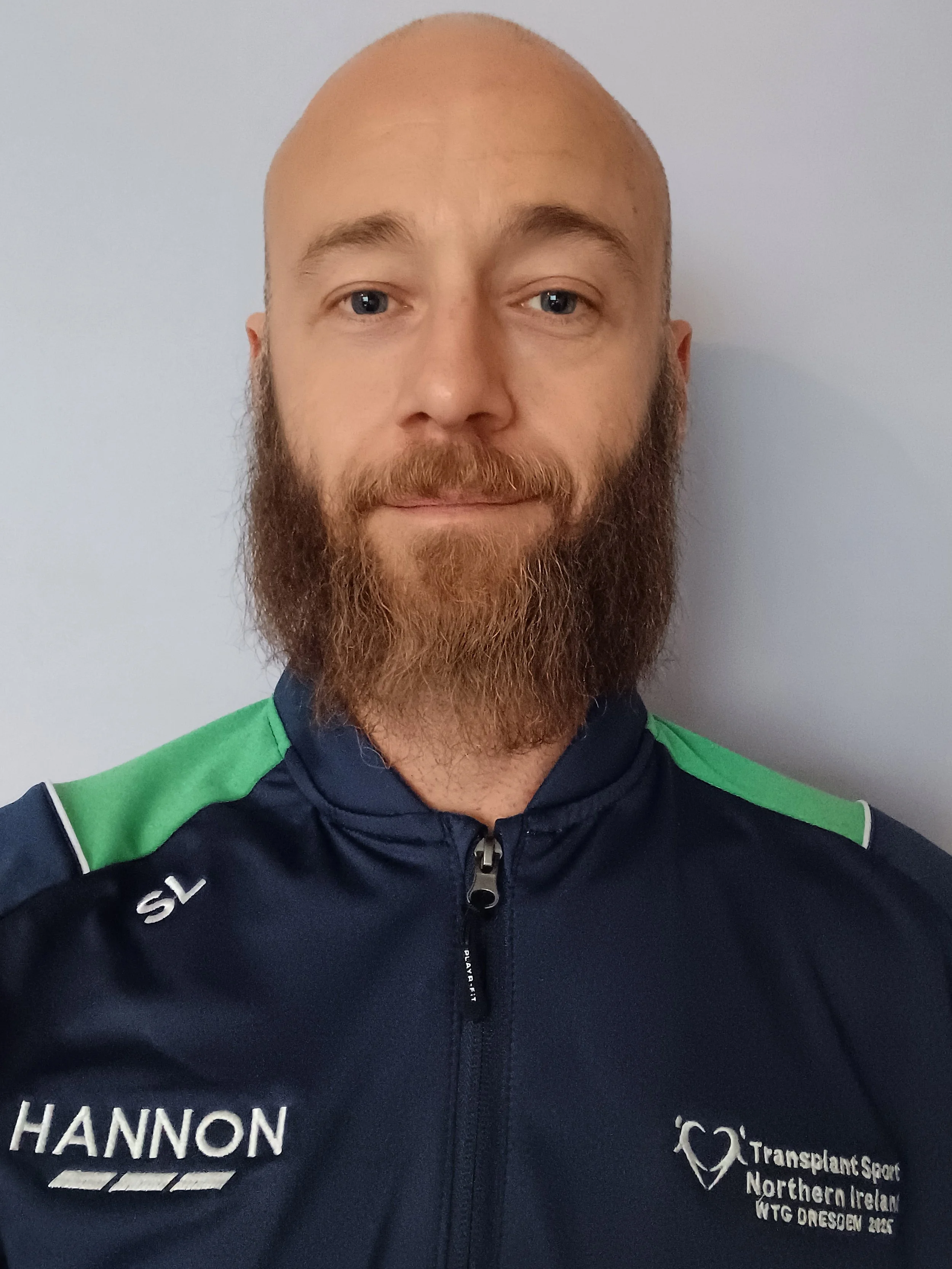 Close-up of a man with a bald head and full beard wearing a sports jacket with logos from Transplant Sport Northern Ireland and the WTG Oresoen 2019, against a plain background.