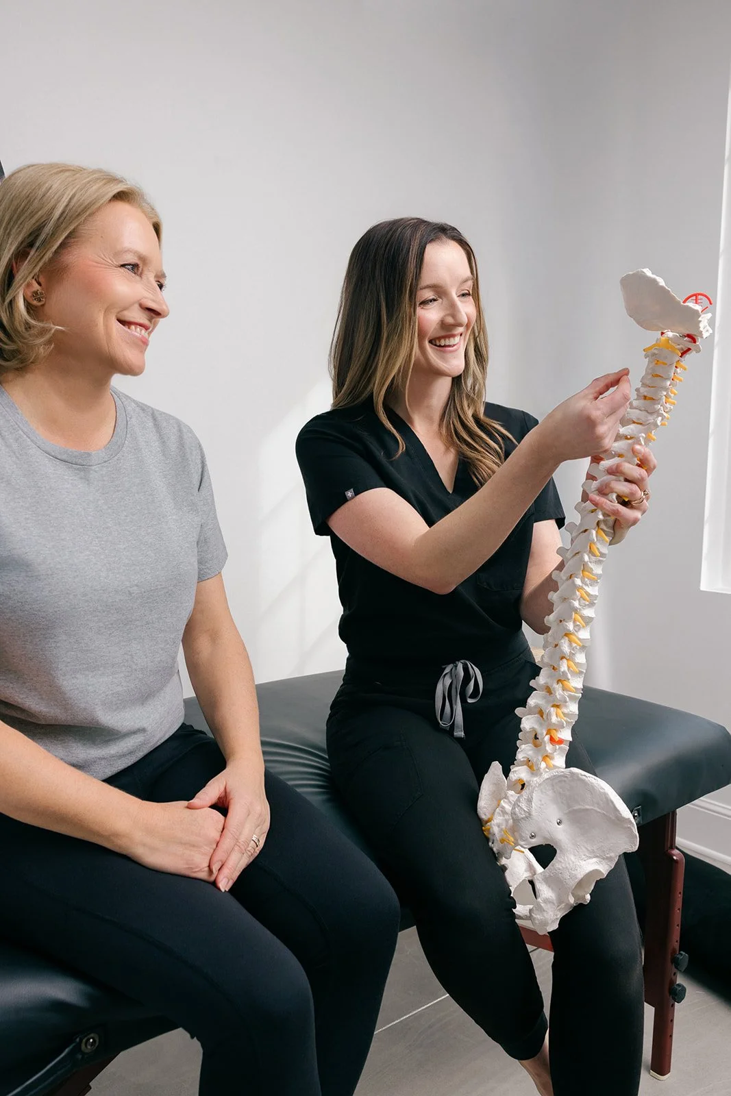 Two women sitting on a medical examination table, one holding a model of a human spine, smiling during a discussion in a clinical setting.