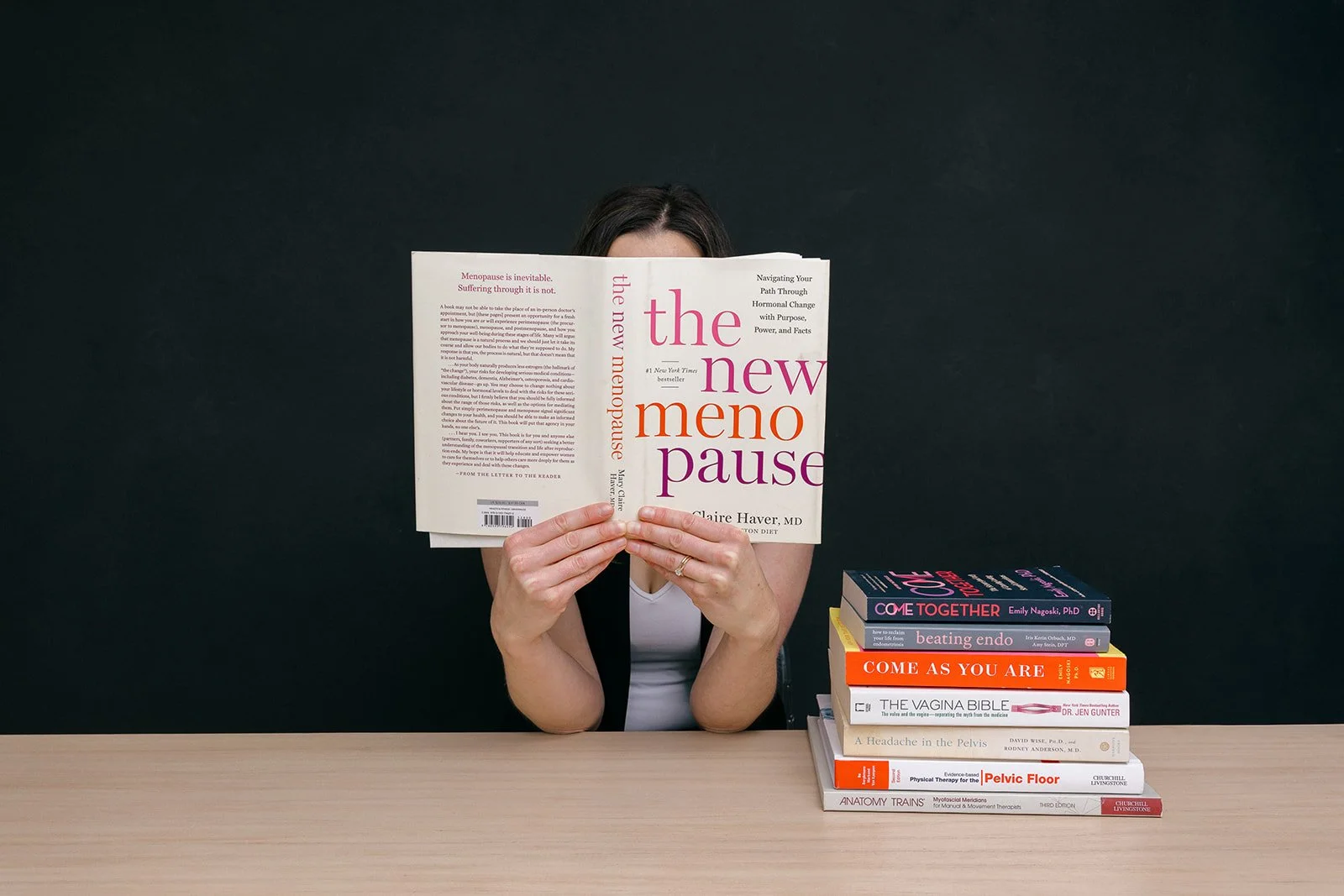 A woman is sitting at a table holding a book titled 'The New Menopause' in front of her face. There is a stack of books on the table beside her.