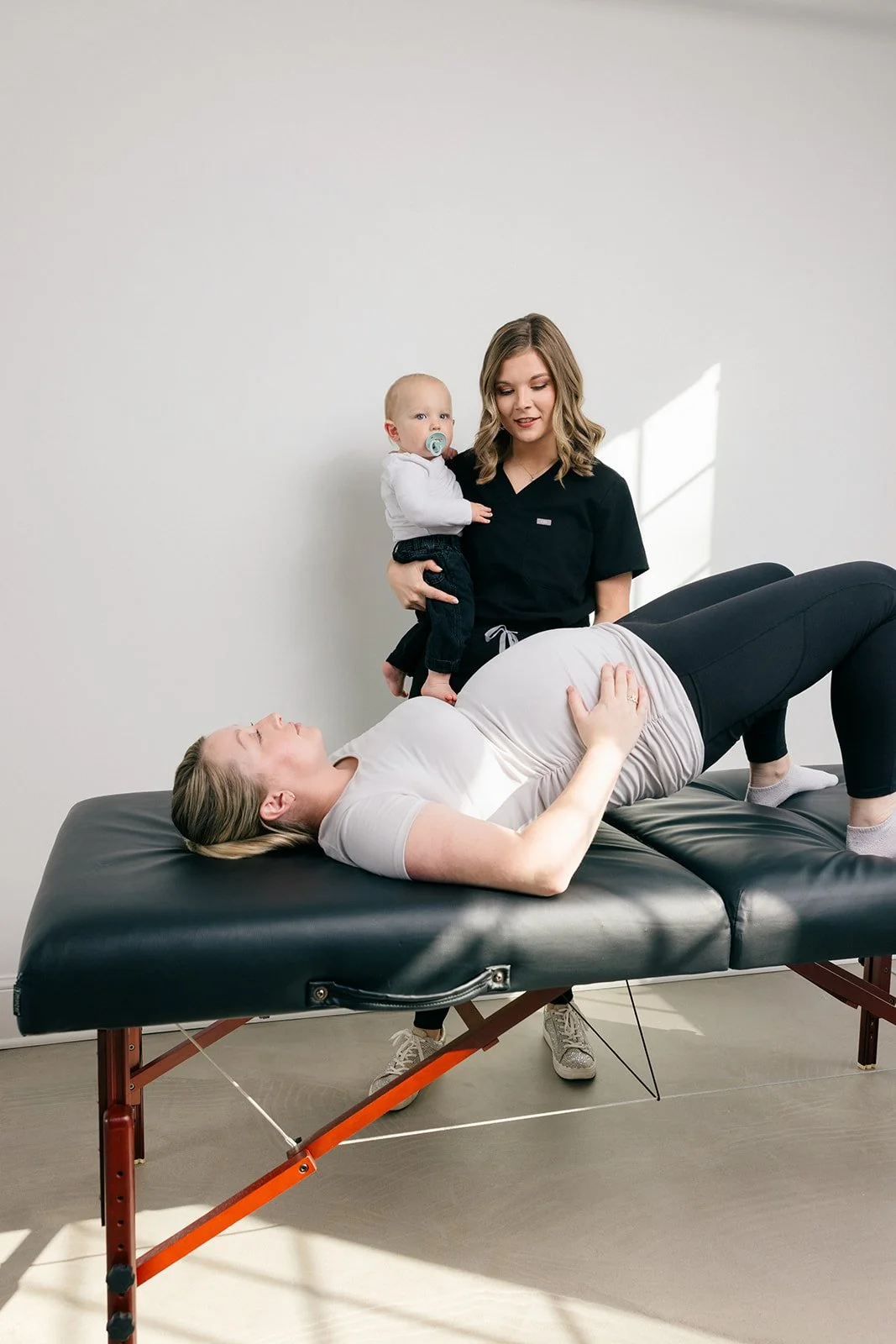 A woman lying on a physical therapy table, a woman holding a baby and a woman lying on the table, likely during a physical therapy osession in a clinic with a plain white wall and natural light.
