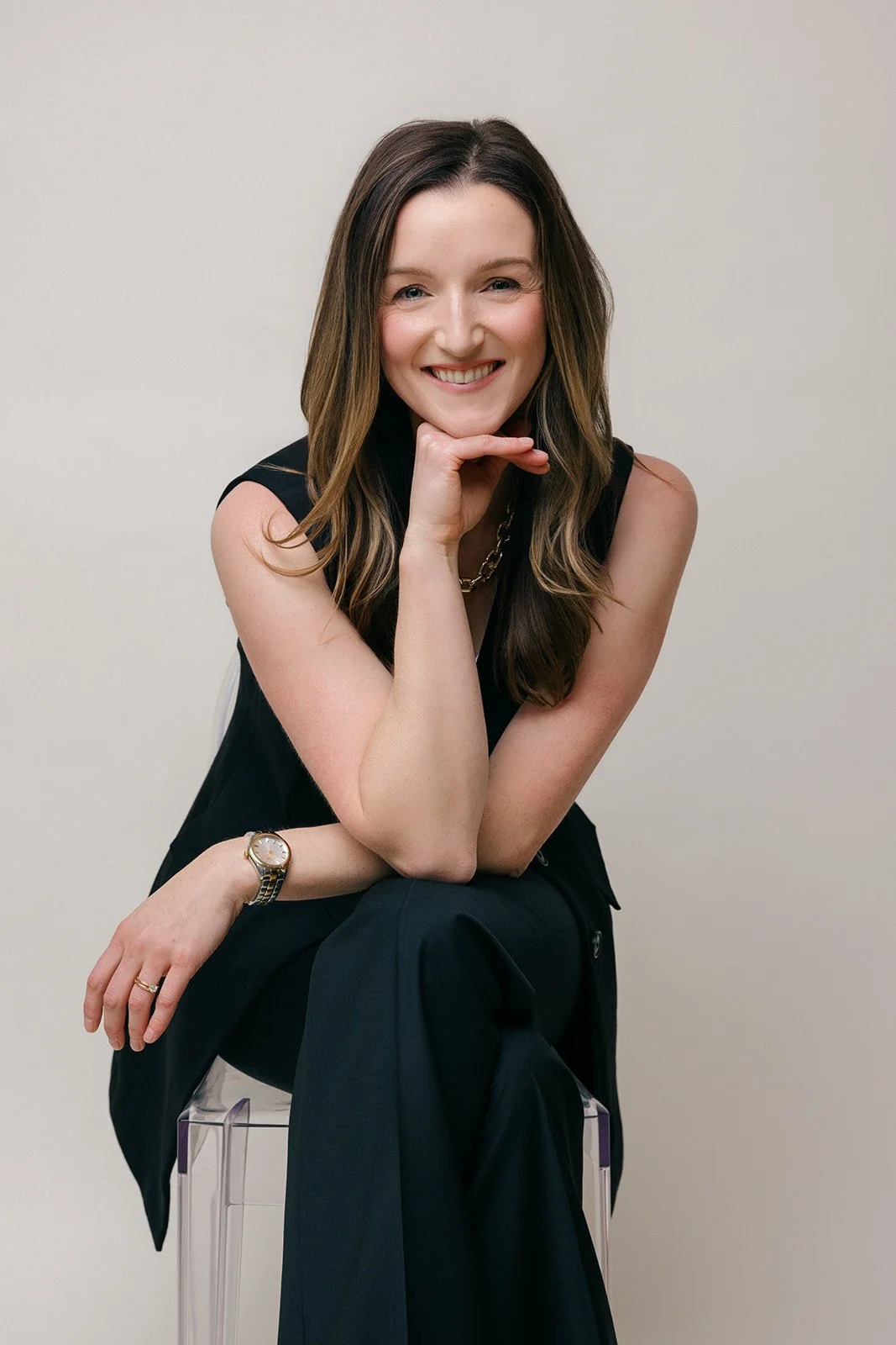 with long, wavy brown hair and fair skin, sitting on a transparent chair against a plain off-white background, smiling and resting her chin on her left hand, wearing a sleeveless black top, black pants, a gold watch, a gold chain necklace, and a ring on her right hand.