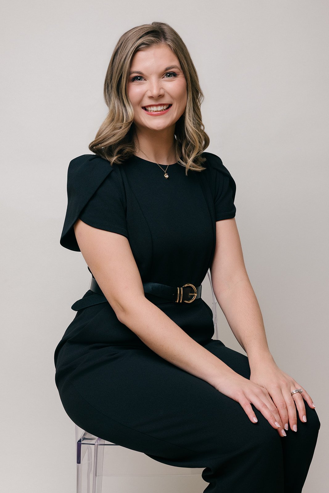A woman with shoulder-length wavy blond hair, wearing a black dress with short puffed sleeves, a black belt with a gold buckle, and a small necklace, sitting on a transparent chair against a plain light background, smiling at the camera.