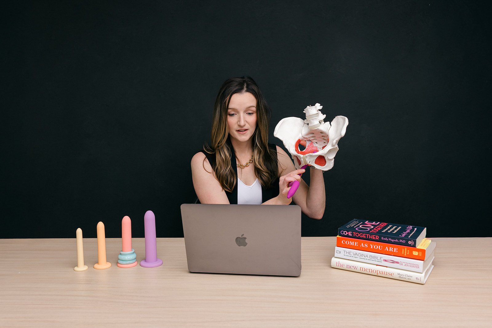 Woman sitting at table with laptop, holding a pelvic bone model, with books about women's health and gender topics, and menstrual cups on the table.