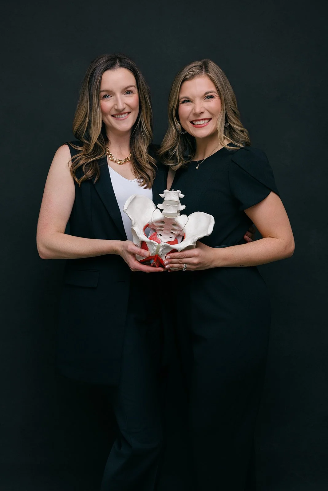 Dr. Emily Priestas and Dr. Meghan Clouspy smiling and holding a pelvic organ model against a black background.