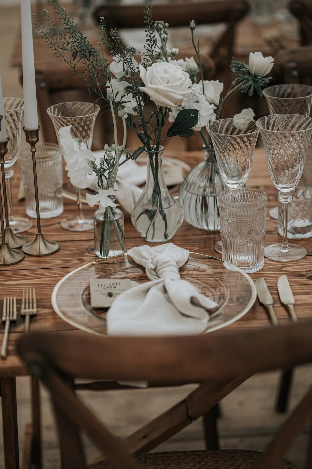A well-decorated dining table with a vase of white flowers, multiple glassware, candles, a plate with a napkin, and gold utensils, set in a rustic wooden setting.