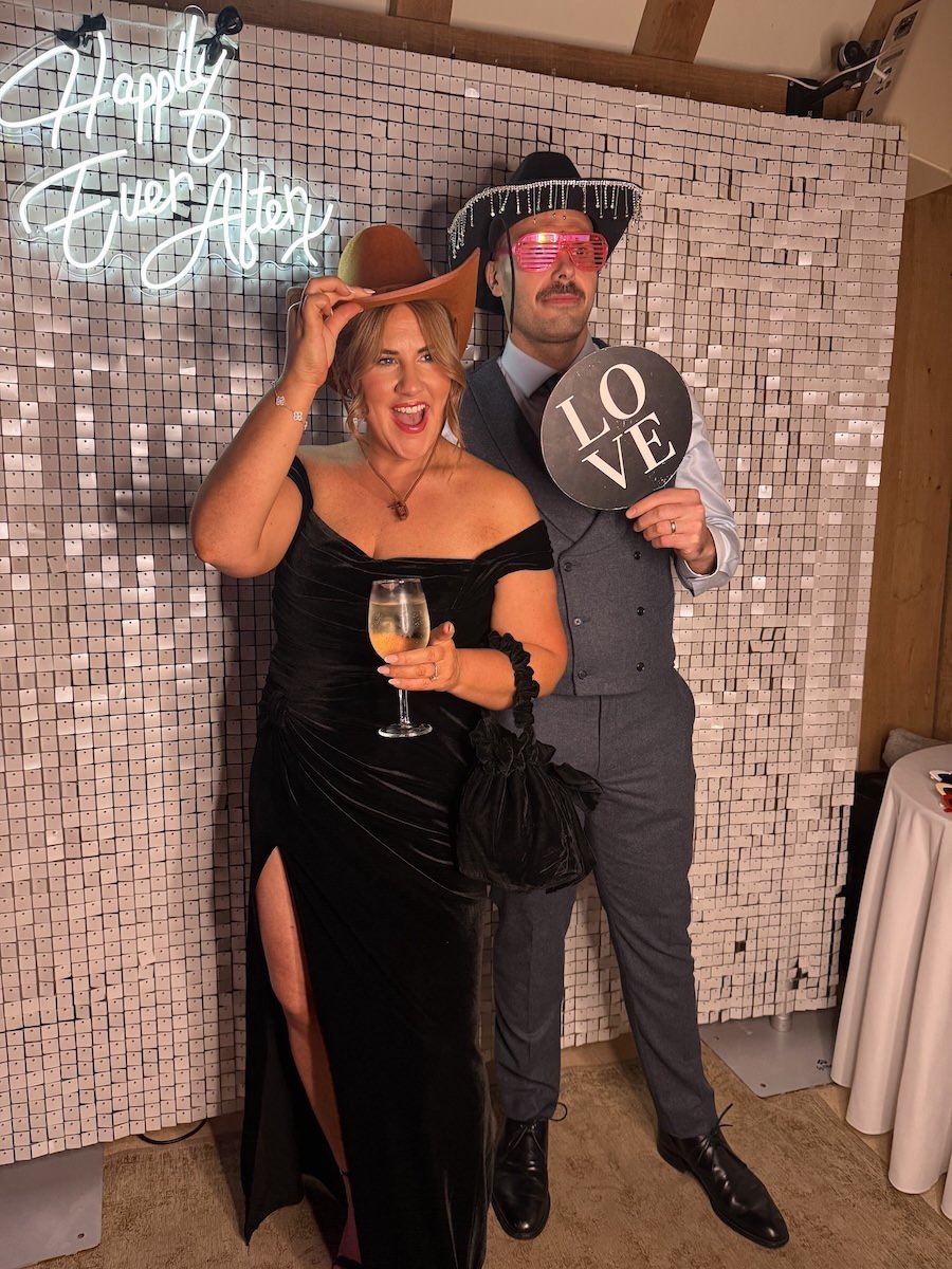 A woman in a black dress holding a glass of wine, smiling, and wearing a large cowboy hat, standing next to a man in a suit with a vest, wearing novelty sunglasses and a sombrero, holding a sign that says "LOVE," at a celebration with a "Happy New Ye