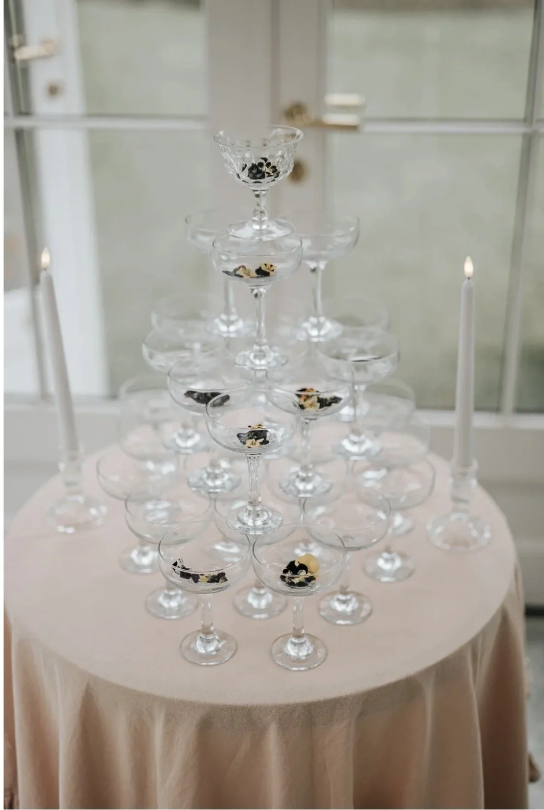 A pyramid of empty cocktail glasses filled with black and white decorative pieces, arranged on a pink tablecloth with two white candles on either side.