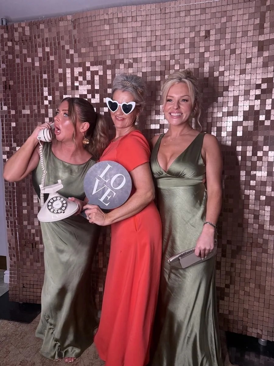 Three women in glamorous dresses posing against a mosaic tile wall. The woman on the left is talking on a vintage rotary phone, the woman in the middle is holding a sign that says 'LOVE' and wearing heart-shaped sunglasses, and the woman on the right