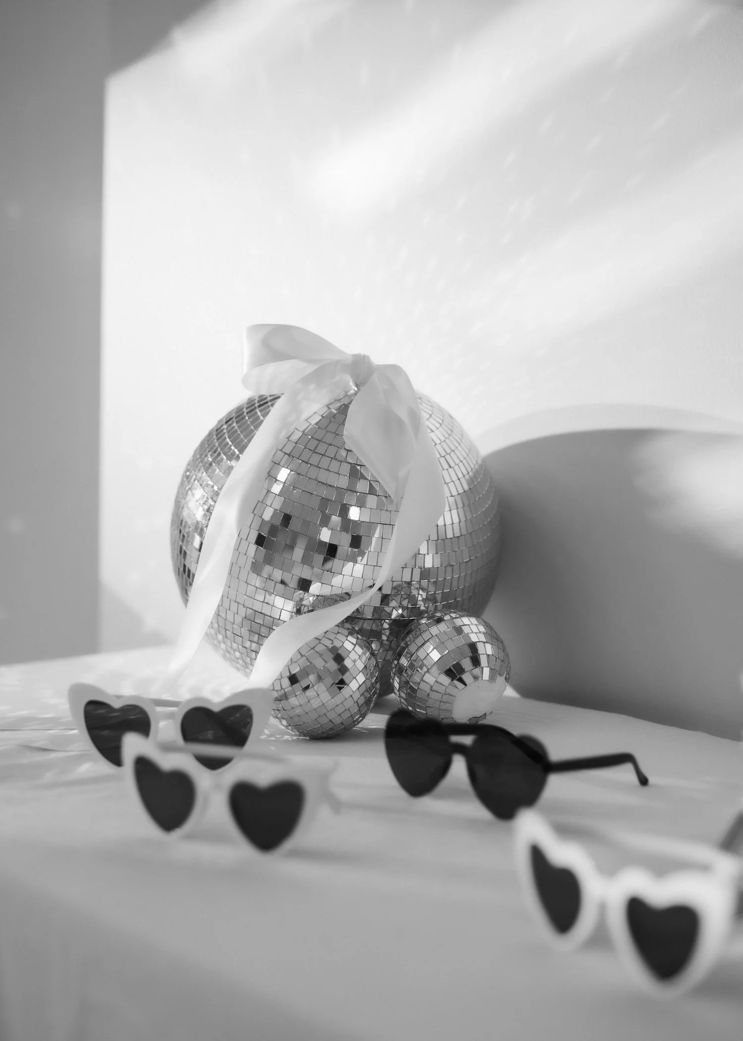 Black and white photo of a large disco ball, with smaller disco balls arranged in front of it, decorated with a white ribbon. Sunglasses are partially visible in the foreground. The setting appears to be decorative or for a celebration.