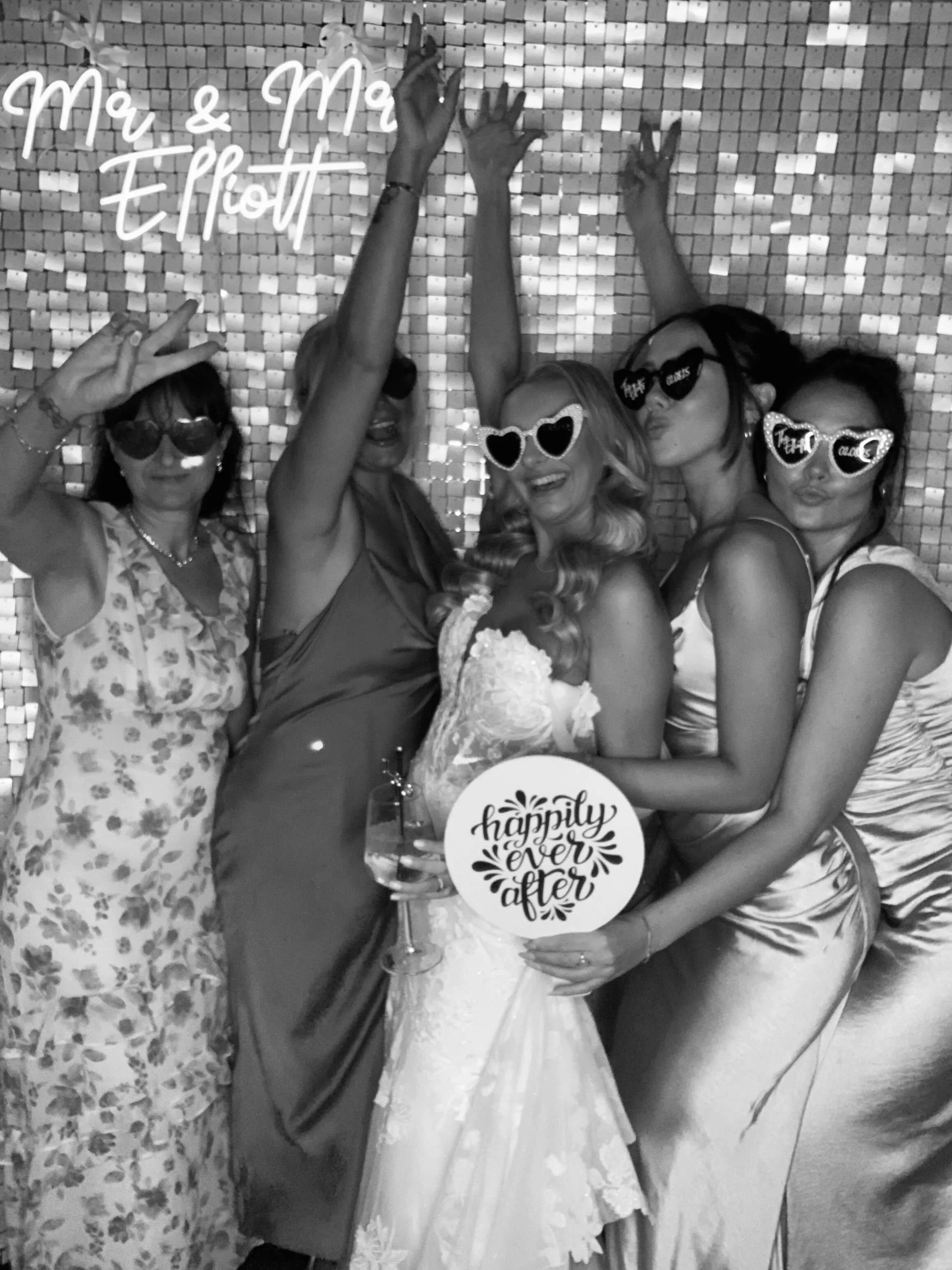 Four women at a wedding reception wearing sunglasses, with a woman in wedding dress in the center holding a sign that says 'happily ever after.' The background has a shiny tiled wall and a neon sign that says 'Mr. & Mrs. Elliott.'