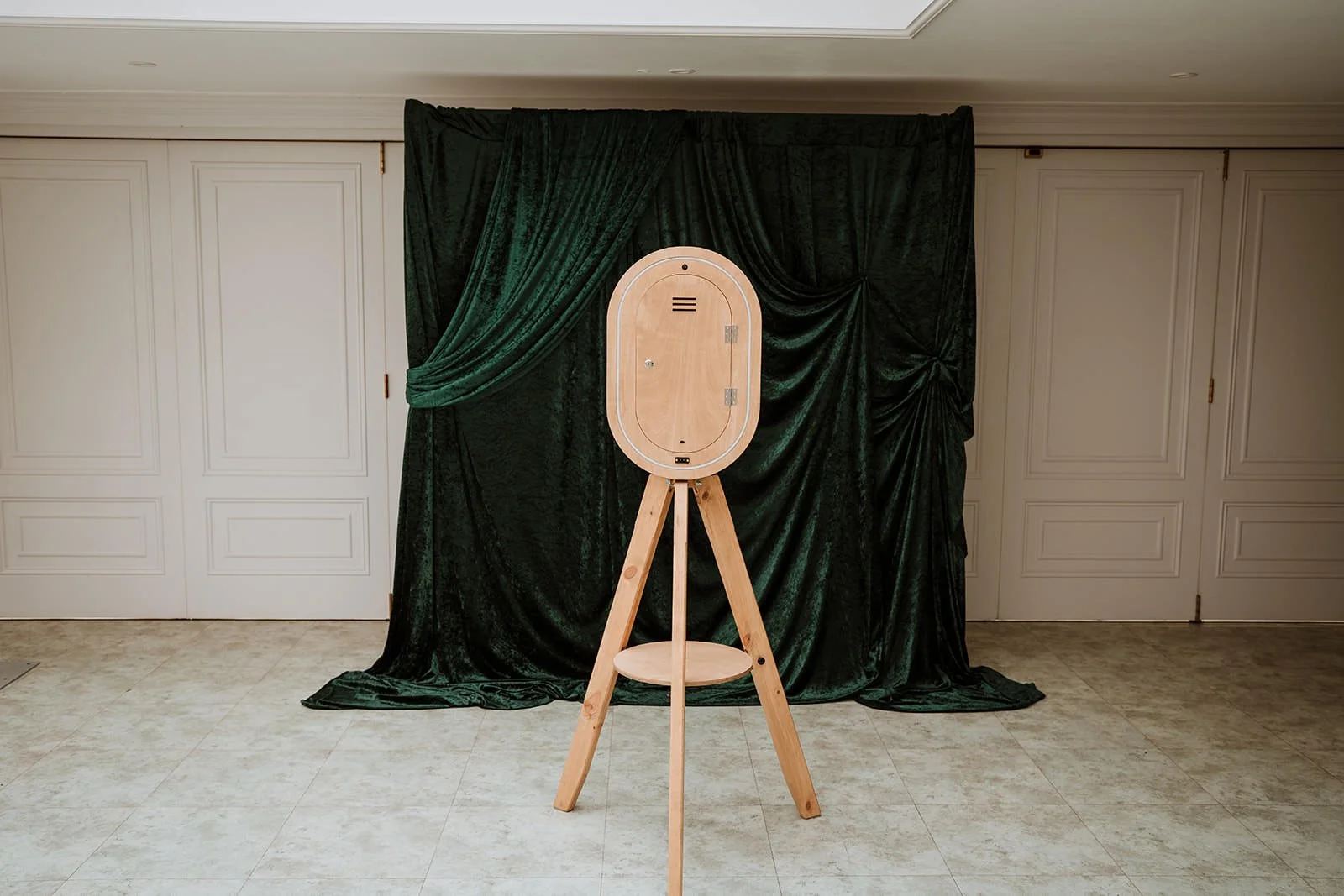 A photo of a wooden photo booth with a tripod stand against a dark green curtain backdrop in an indoor setting.