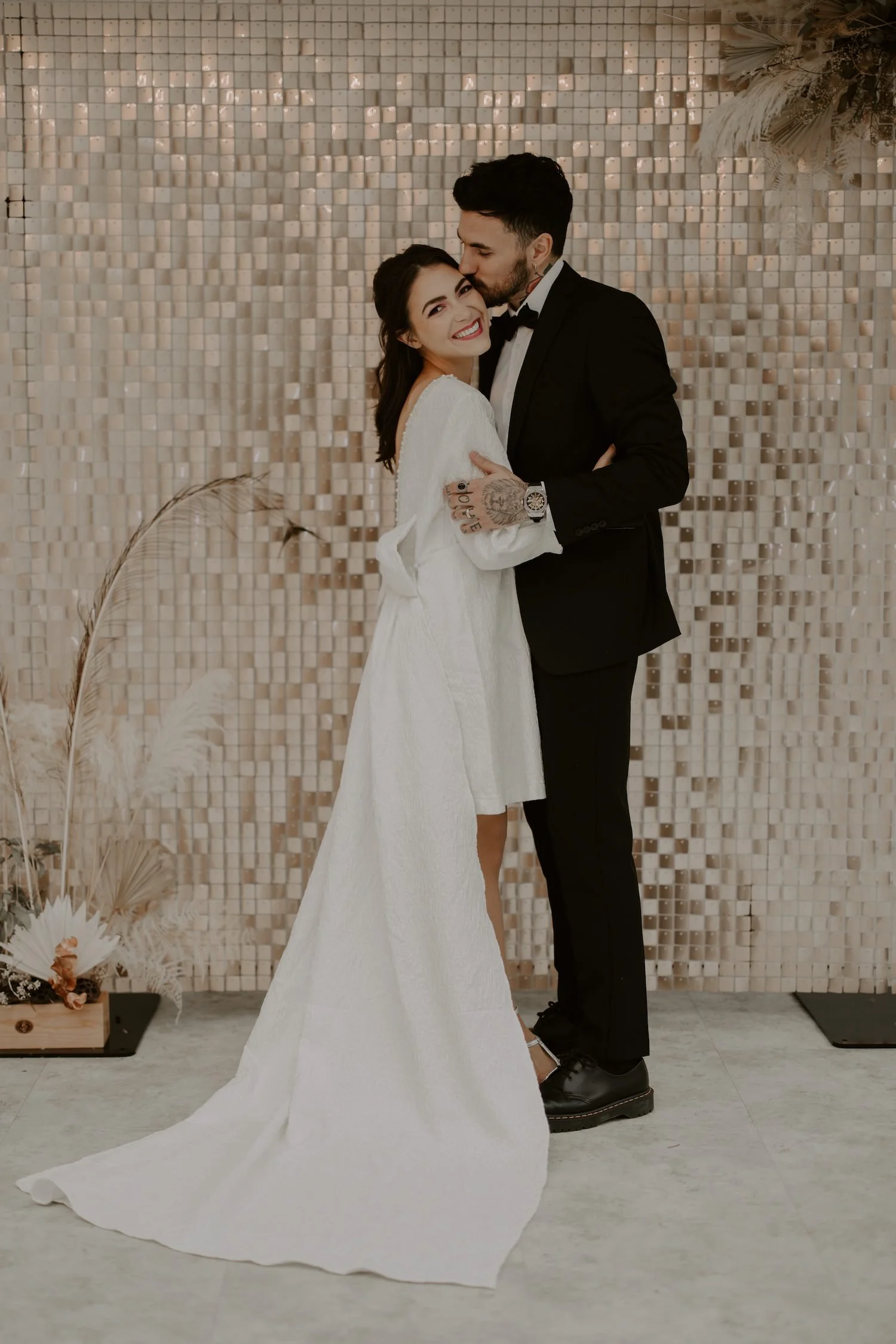 A bride and groom dancing together at their wedding reception. The bride is smiling happily, wearing a white wedding dress, and the groom is dressed in a black suit and bow tie, leaning in to kiss her forehead. The background features a decorative wa