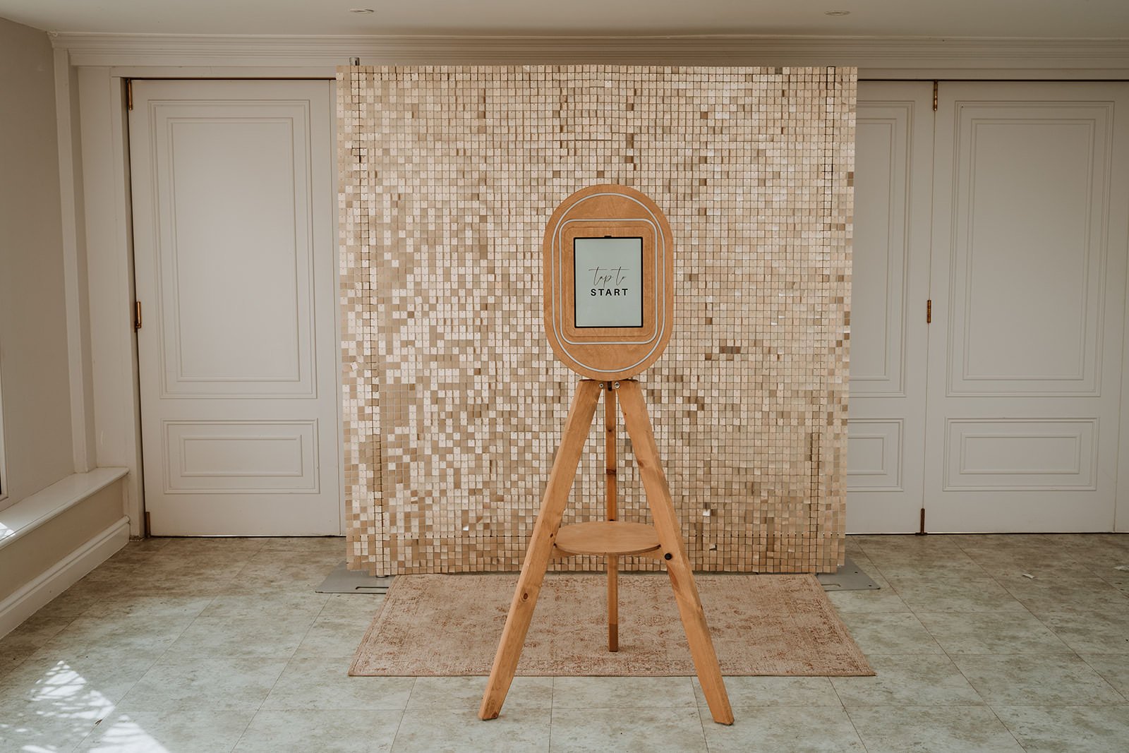 An indoor setup with a wooden stand holding a digital device displaying the words "top to START". The background features a decorative wall with small, shiny, beige tiles, and the floor has light-colored tiles with a small beige rug. There are white 
