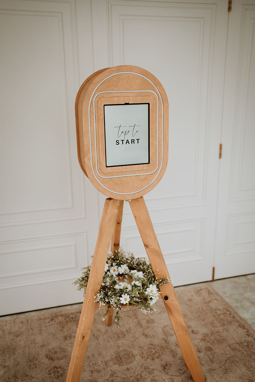 A wooden photo booth with a digital screen displaying "Tap to START" in a room with white paneled doors and beige carpet, decorated with a small flower wreath at the base of the stand.