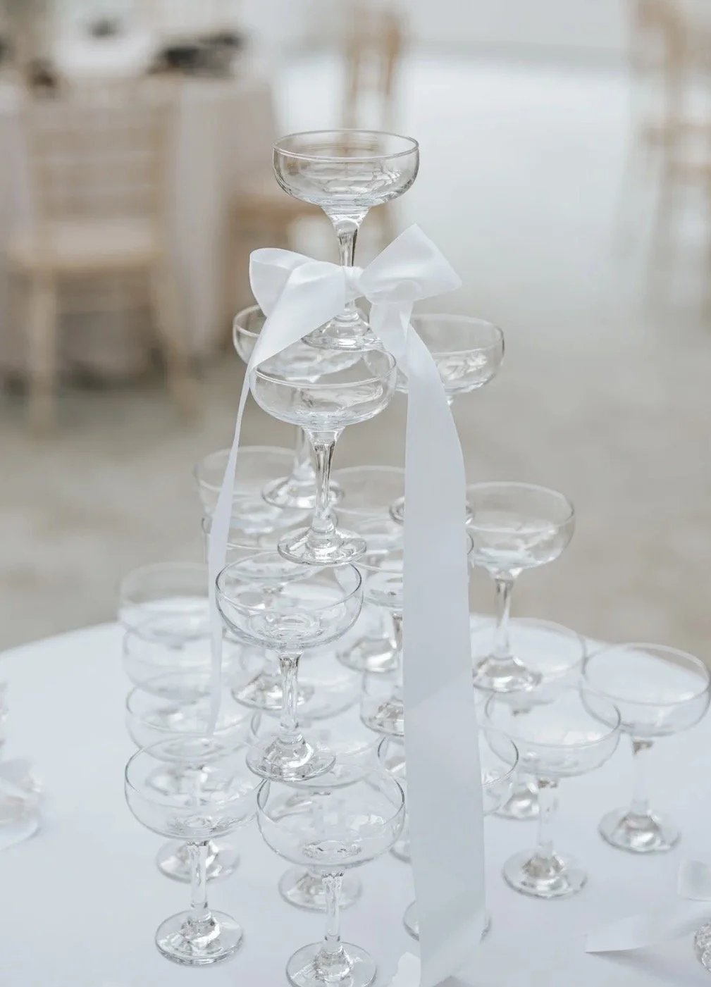 A pyramid of empty champagne glasses decorated with a white ribbon, set on a table at a celebration or wedding event.
