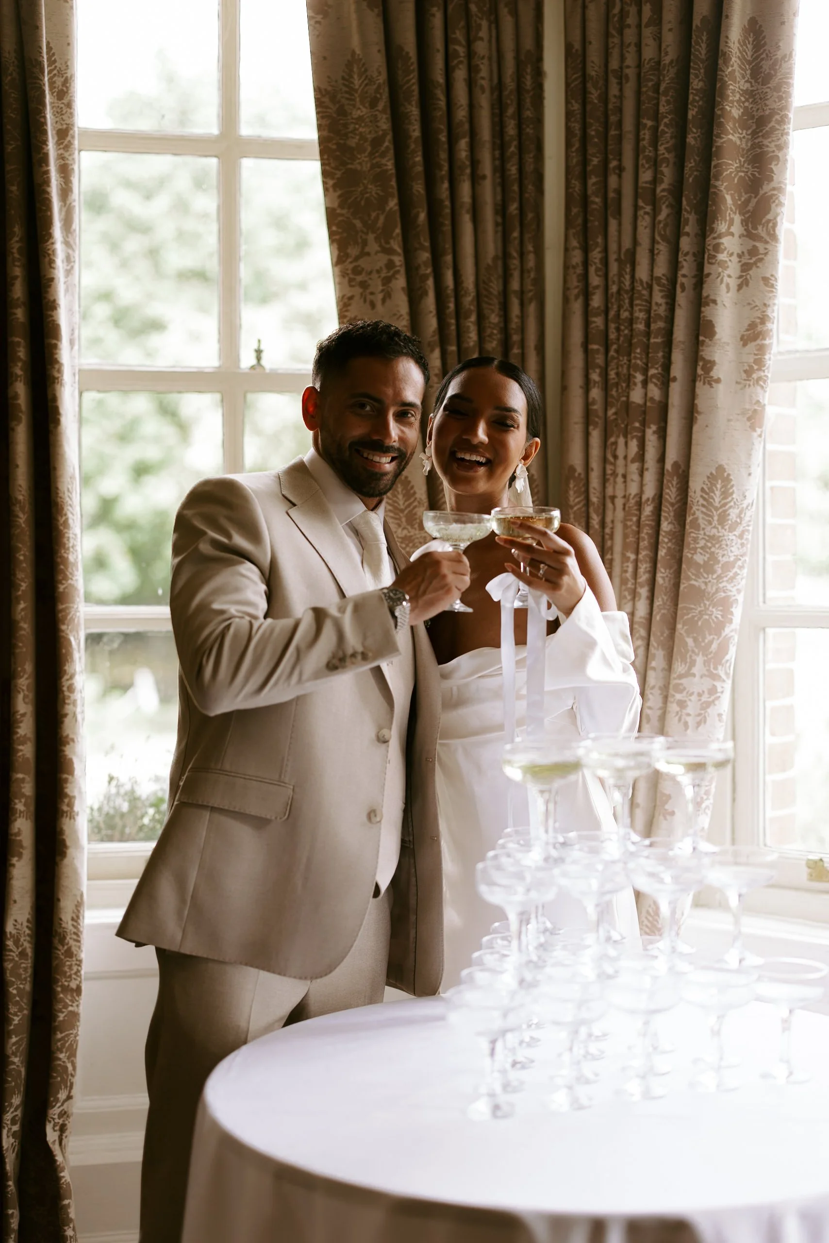 A smiling man and woman dressed in formal attire celebrating with champagne at a wedding reception, standing behind a champagne tower.