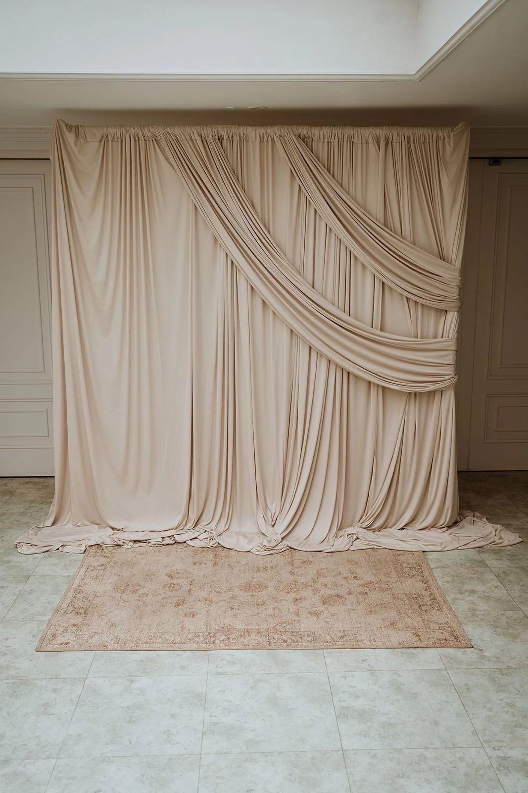 Cream-colored draped backdrop elegantly across a window, hanging above a beige rug on cream tile flooring in a room with white walls and trim.