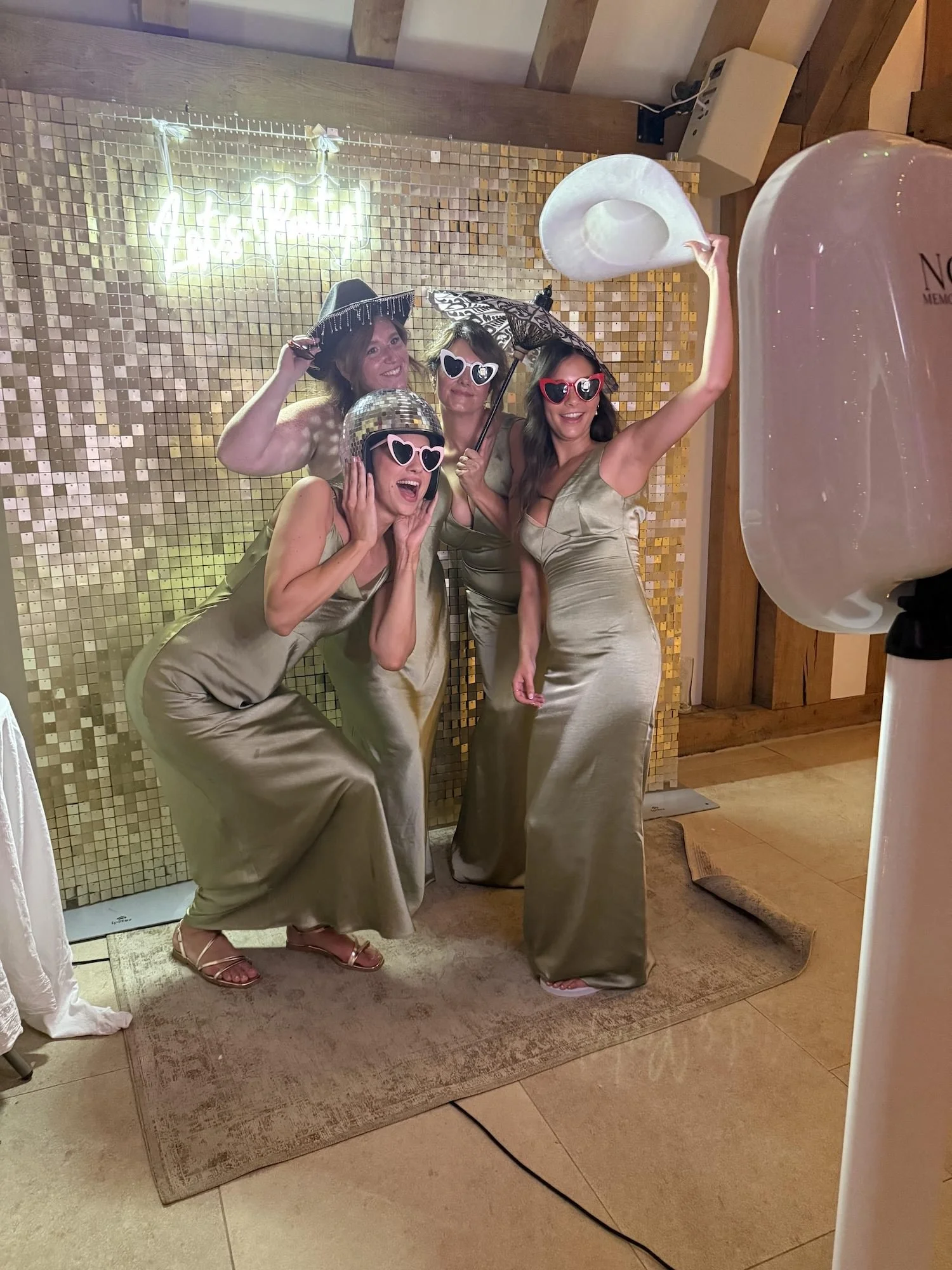 Four women in gold dresses and sunglasses celebrating at a party, posing for a photo with hats and umbrellas, standing on a beige rug in front of a gold tiled wall with illuminated sign in background.