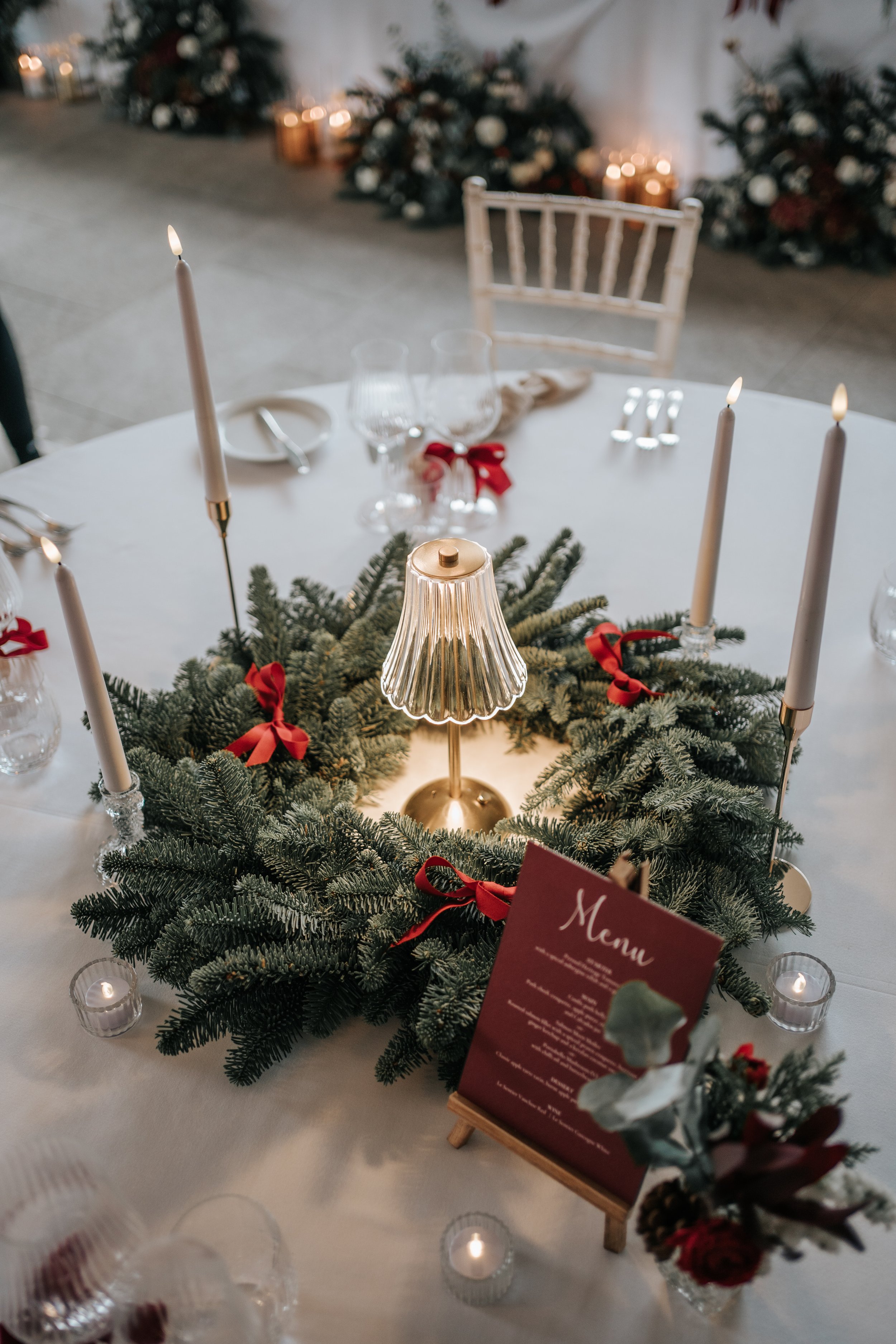 A Christmas table centerpiece with a gold lamp, evergreen branches, red ribbons, lit candles, and a menu card, set on a white tablecloth with glasses and utensils, and blurred Christmas decorations in the background.