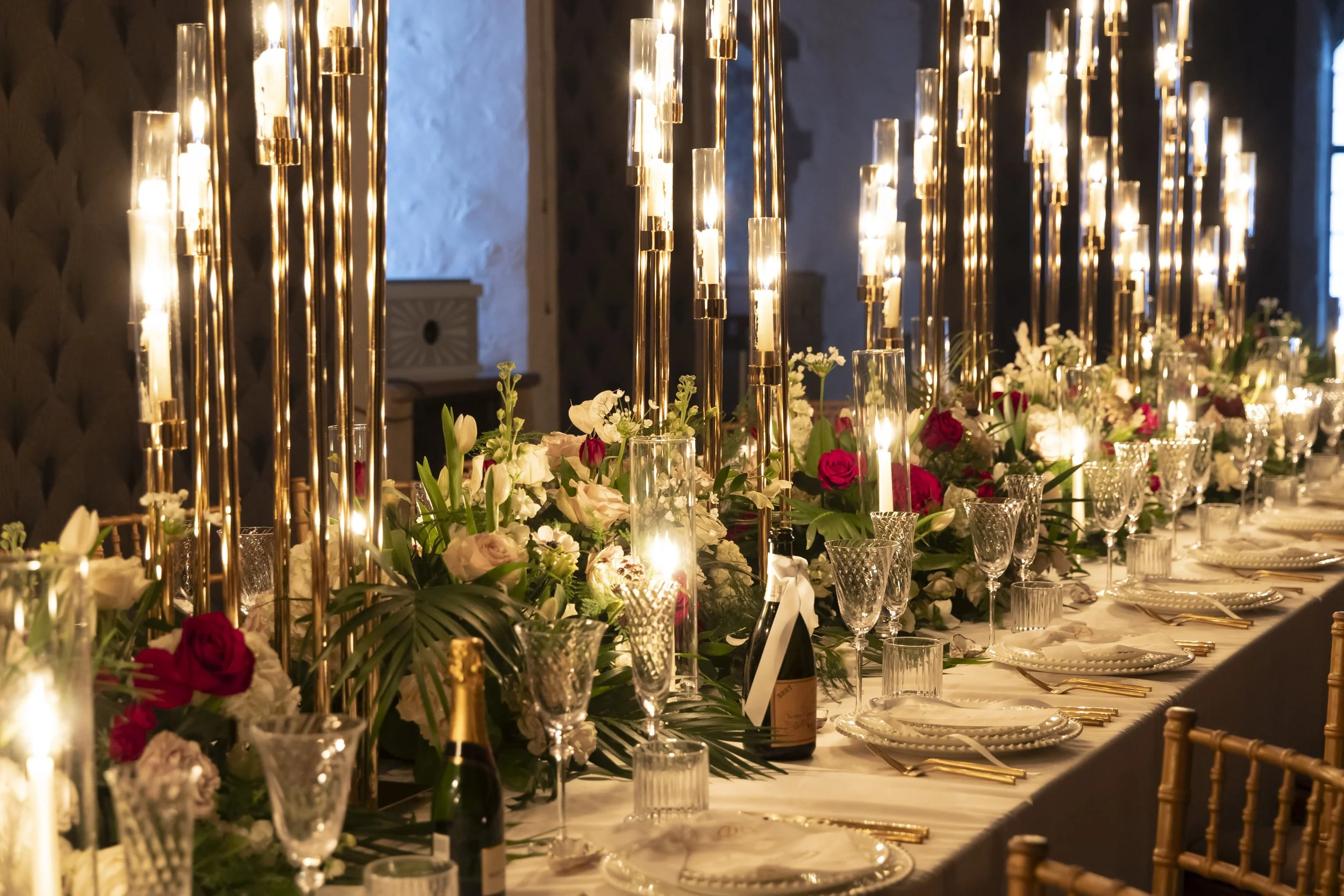 A luxurious banquet table decorated with tall candle holders, floral arrangements with pink and white flowers, and gold accents, set for a formal event in an elegant room with dim lighting.