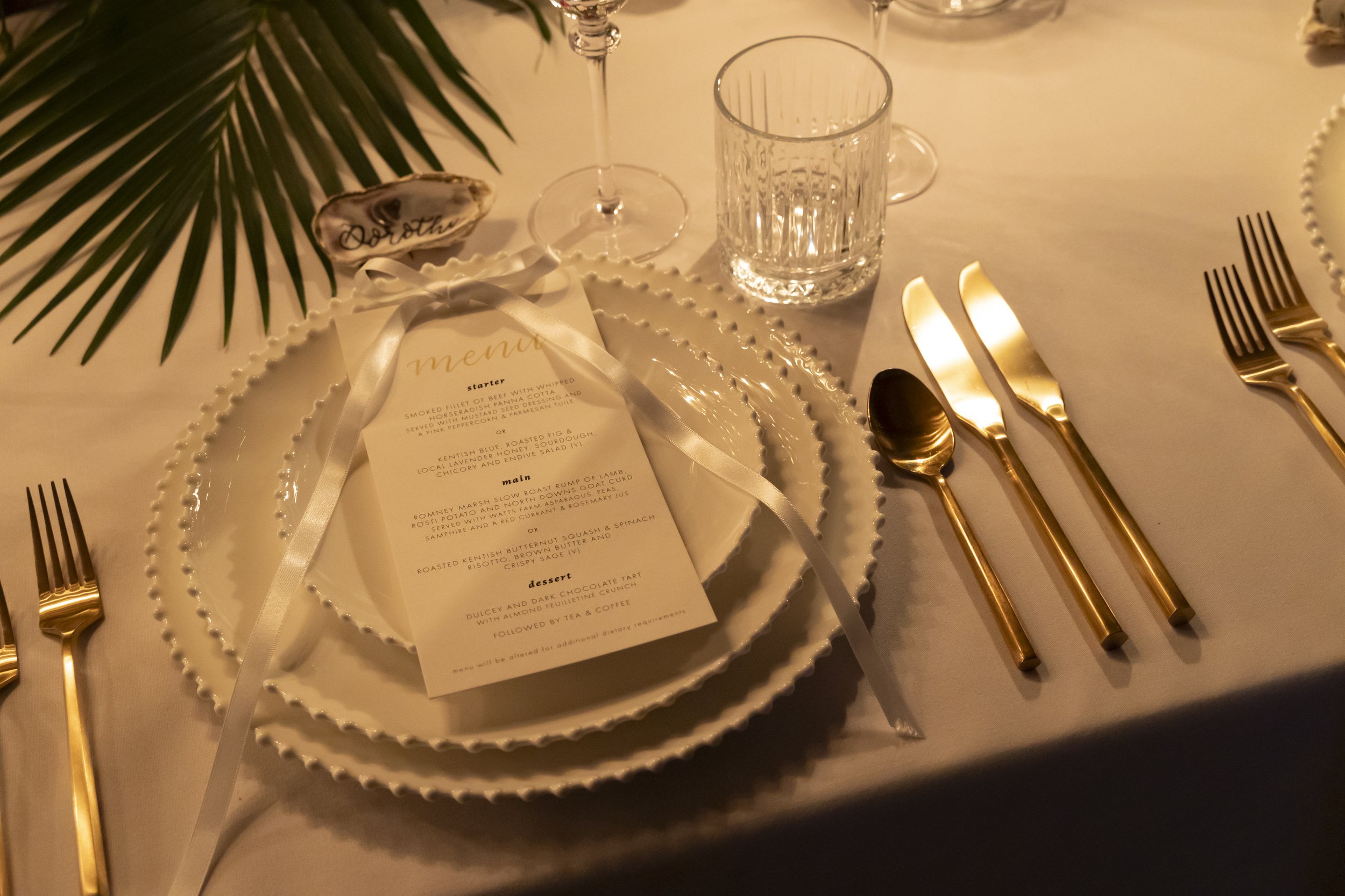 Elegant table setting with a menu, gold flatware, glassware, and decorative elements including a palm leaf and a sign with cursive writing, possibly at a formal event or restaurant.