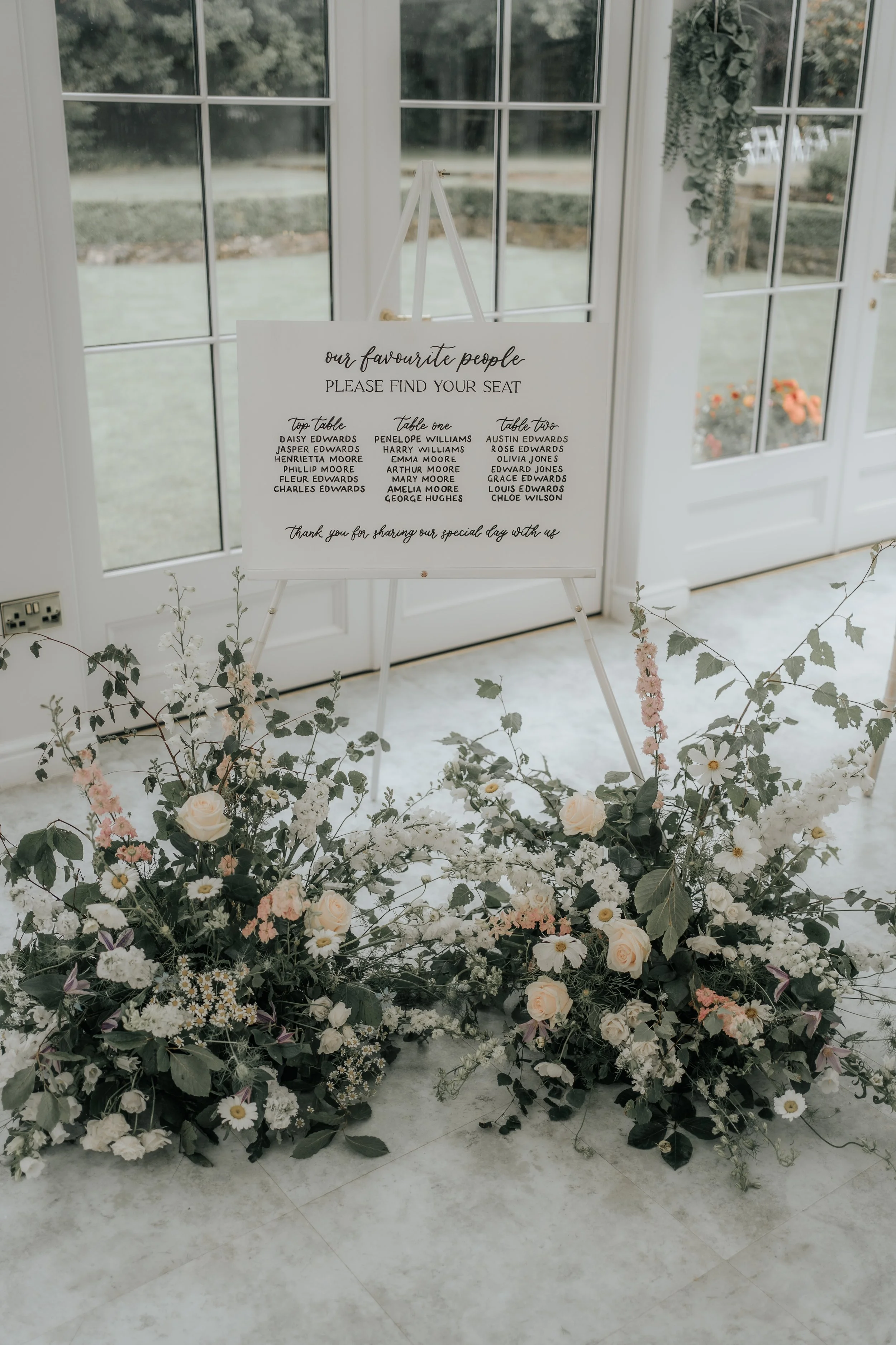 A floral wedding display with a table seating arrangement sign, surrounded by white, pink, and purple flowers and greenery, set inside near glass doors and windows with a garden view outside.