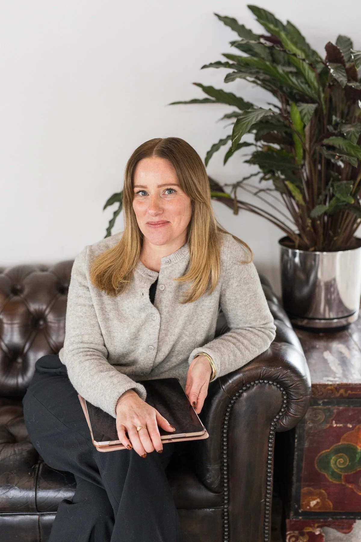 A woman with light brown hair, wearing a gray cardigan, sitting on a brown leather tufted sofa, holding a tablet, with a large potted plant with dark green and purple leaves behind her, against a plain white wall.