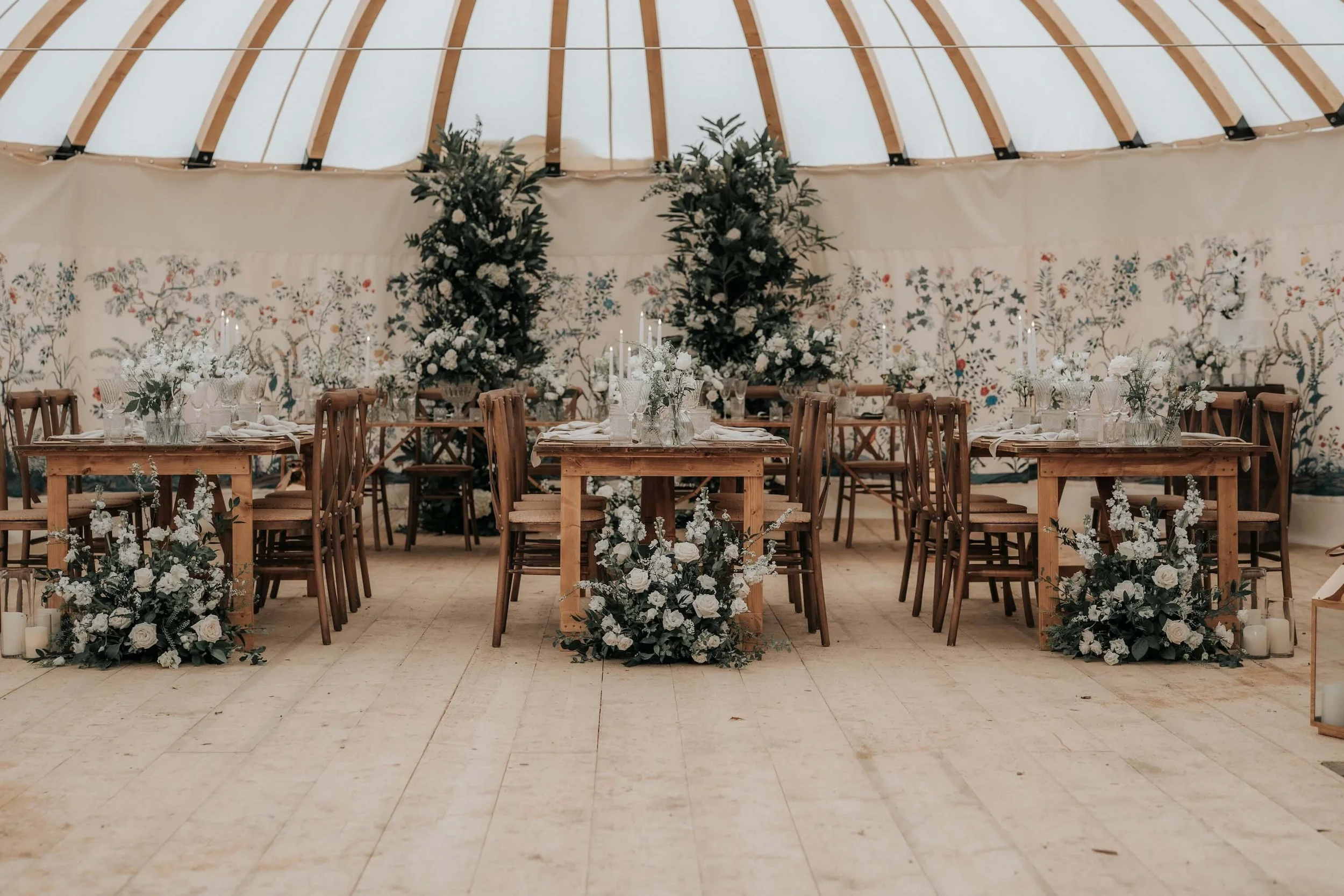 A decorated event space with wooden tables and chairs, floral arrangements, and large greenery and white floral decorations in the background under a partially constructed wooden ceiling.