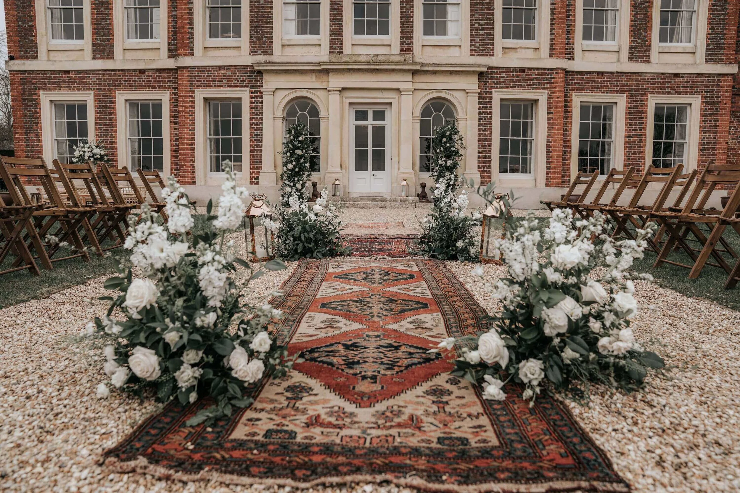 Outdoor wedding setup with wooden chairs arranged on both sides of a red and beige patterned rug, surrounded by white flowers and greenery, in front of a historic brick building.