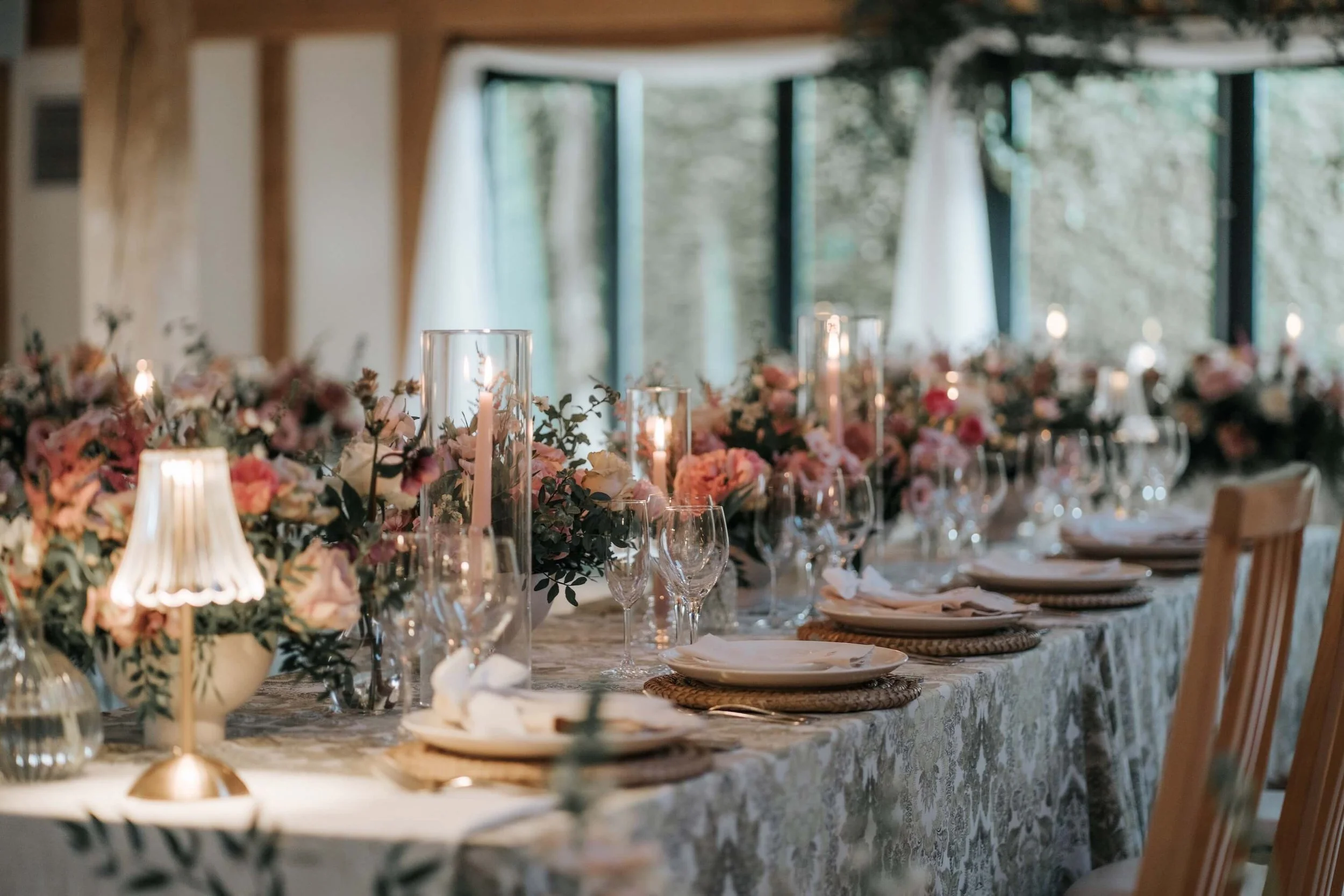 A decorated banquet table with floral centerpieces, candles, wine glasses, and table settings in a well-lit venue with large windows and draped curtains.