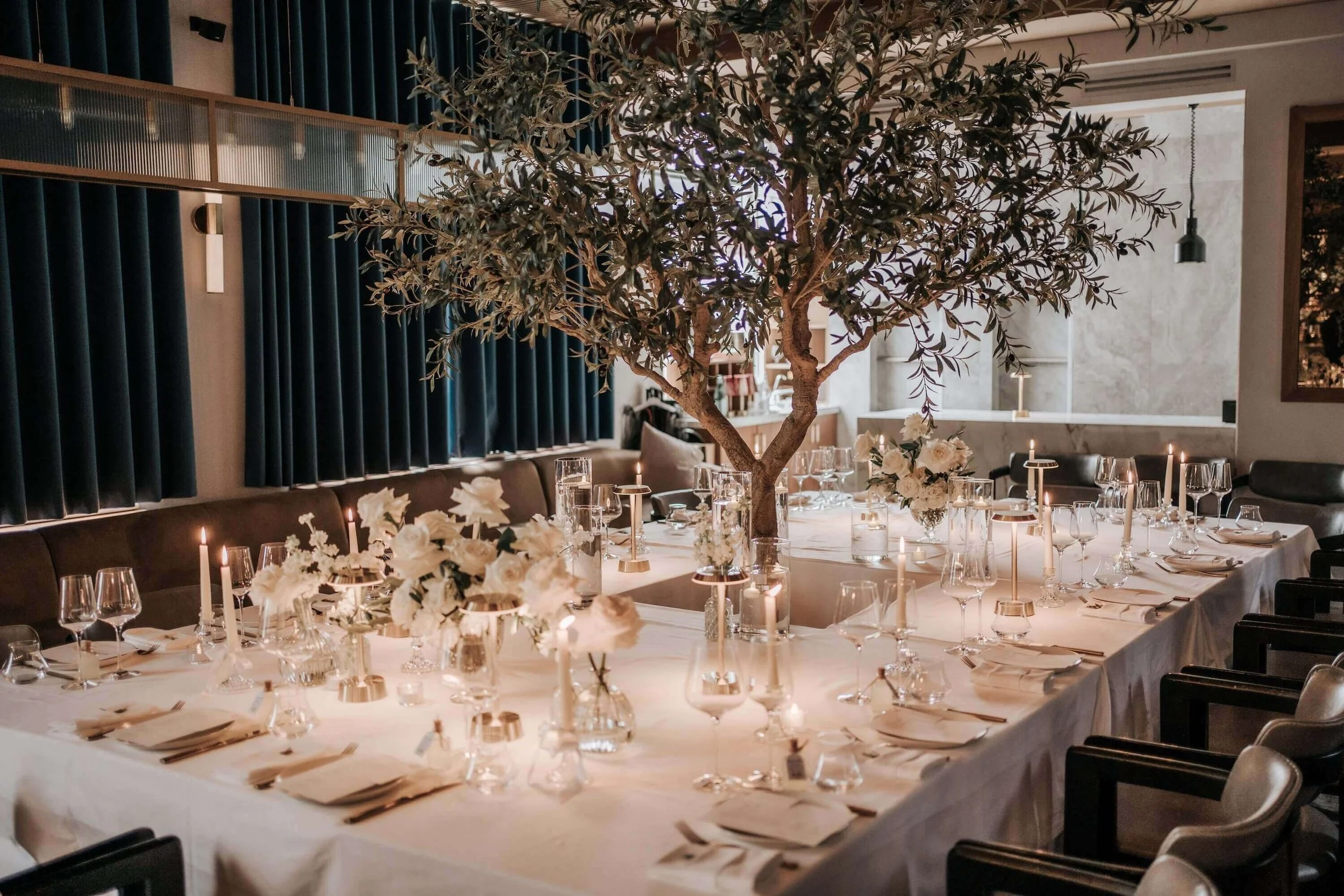 Elegant dining table set for a special event with white floral centerpieces, candles, and glassware, featuring a large olive tree in the center of the room.
