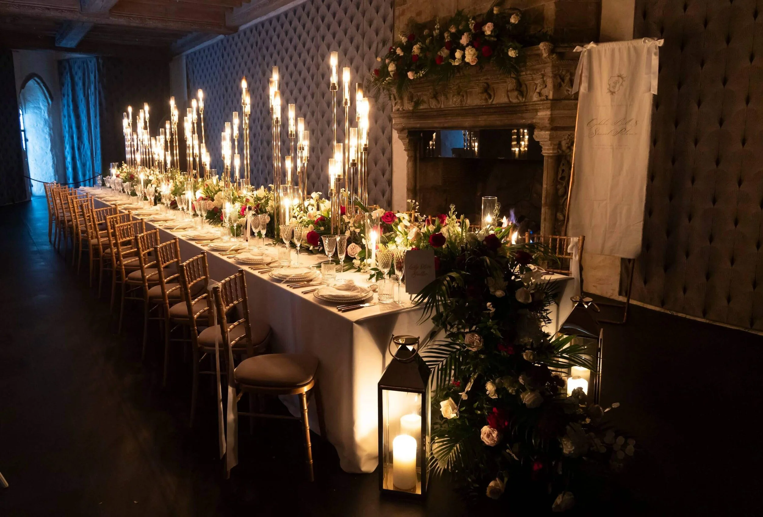 Elegant wedding reception table with tall candles, floral arrangements, gold chairs, and a fireplace with flowers on top in a dimly lit room.