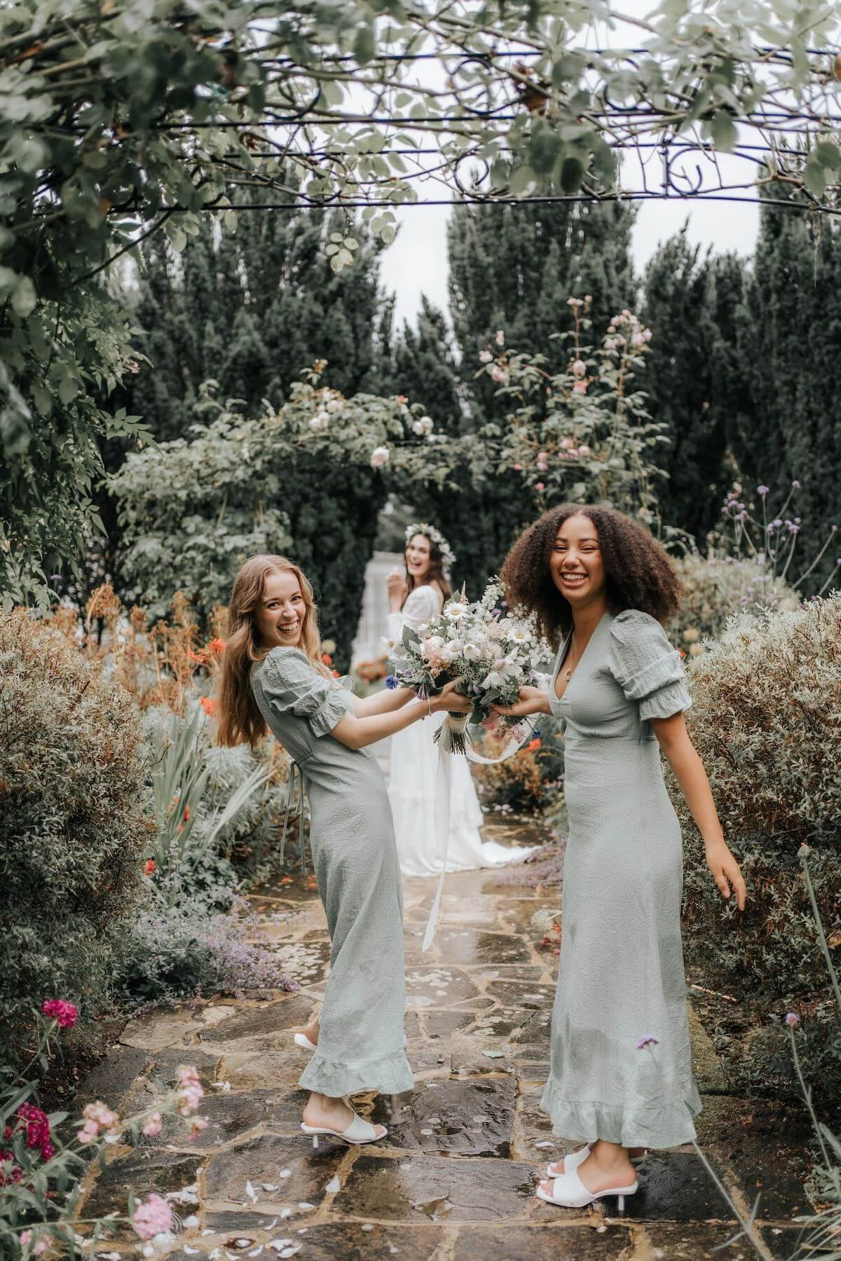 Two women in matching gray dresses holding a bouquet and smiling at each other in a garden, with a woman in a white dress in the background.