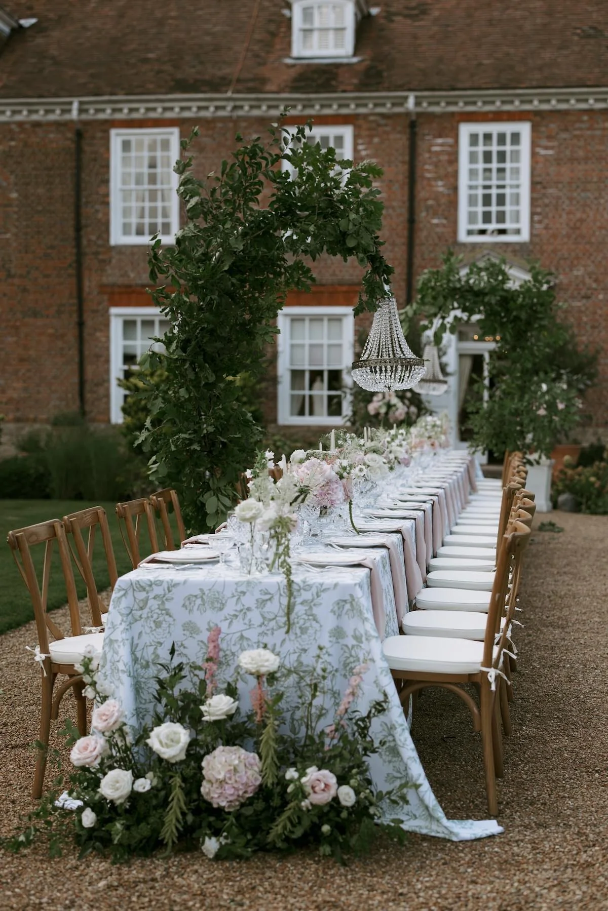 Long outdoor dining table decorated with white floral tablecloth, pink and white flowers, and greenery, set with glassware, surrounded by wooden chairs with white cushions, under a chandelier, in a garden courtyard near a brick building with white-fr