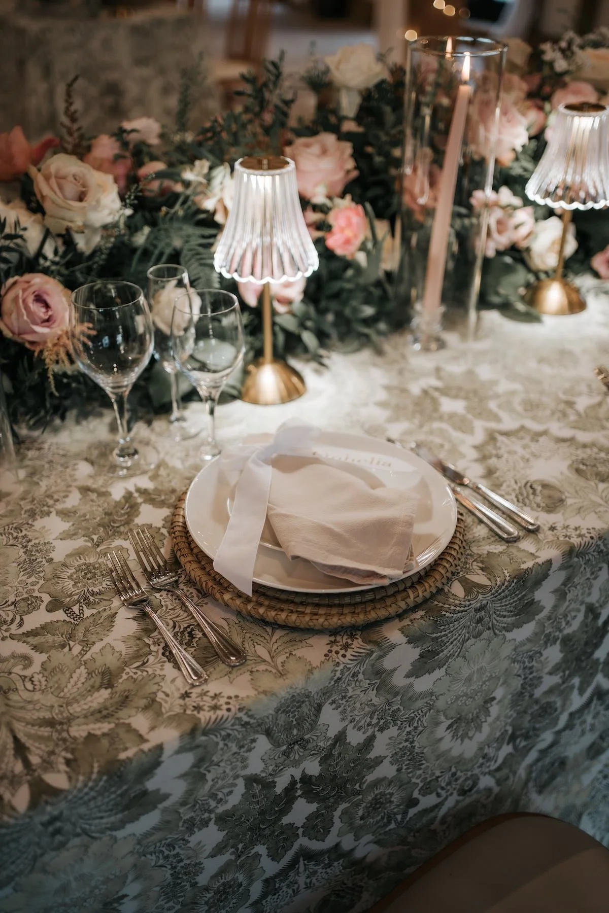 Elegant dinner table setting with floral centerpiece, glasses, cutlery, and a napkin on a patterned tablecloth, illuminated by small lamps.