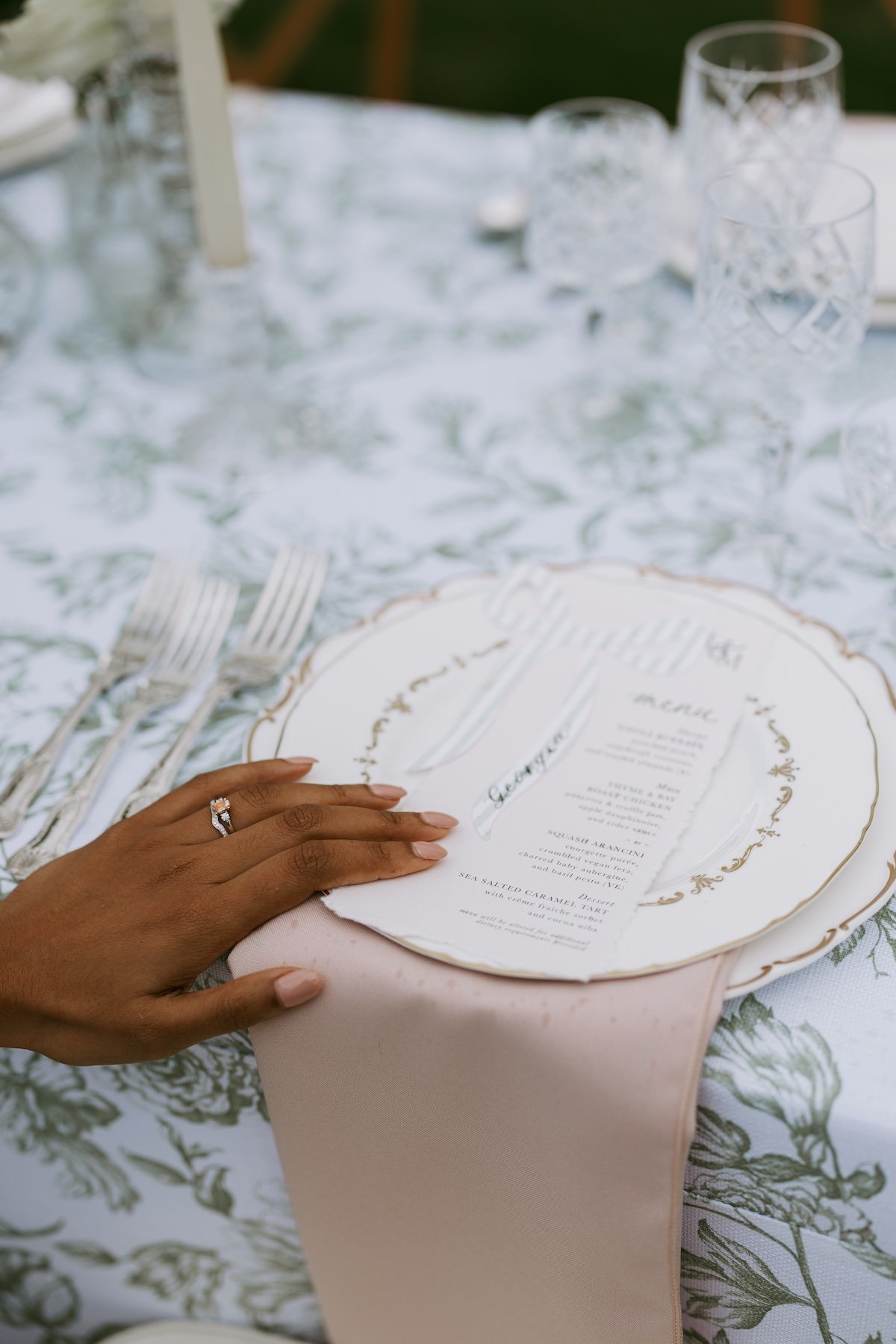 Close-up of a person's hand with a ring, resting on a place setting with a printed menu on a decorative plate, at an outdoor table with a floral tablecloth and glassware in the background.