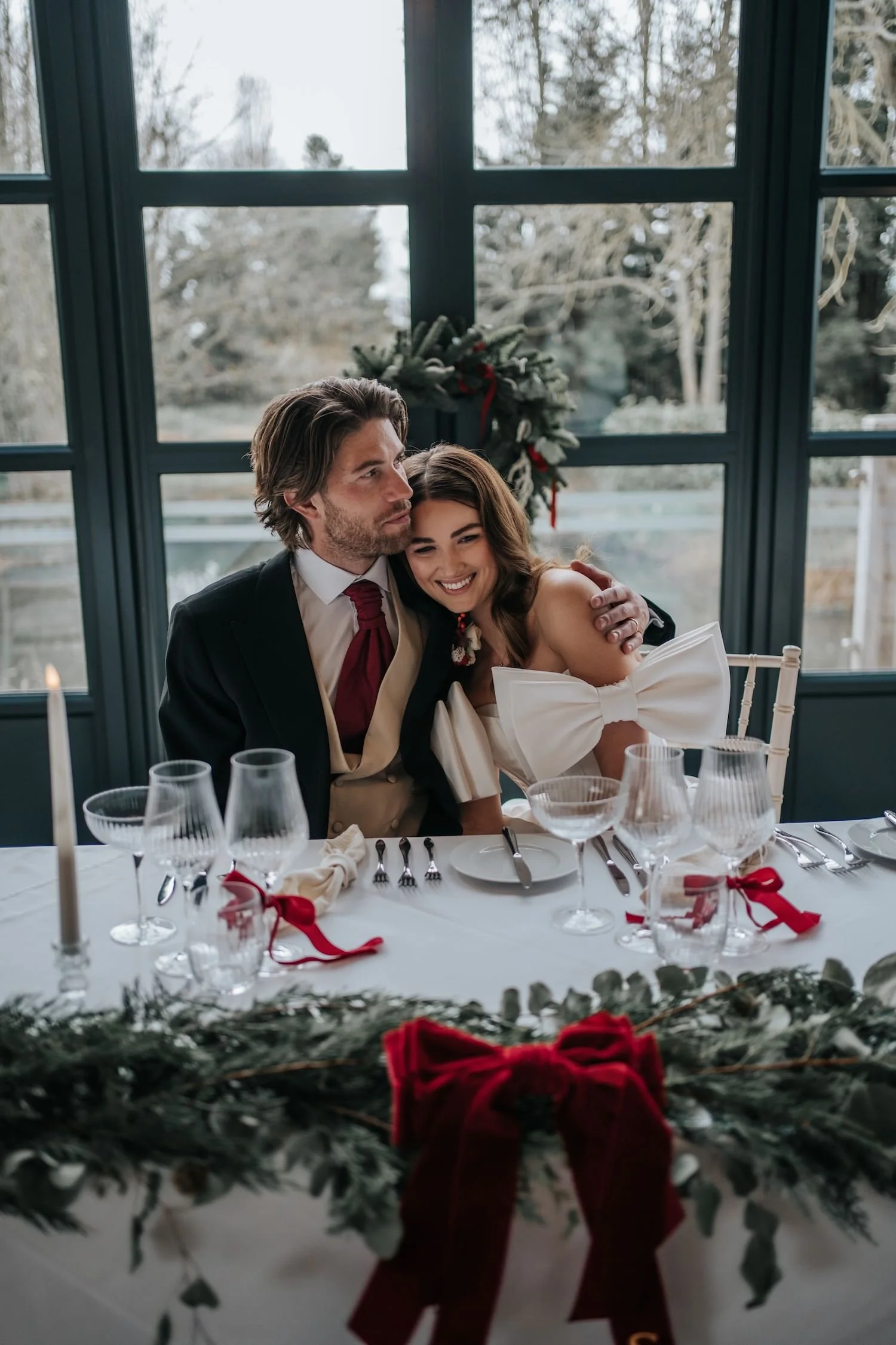 A man and a woman sitting close together at a dining table, smiling and embracing each other, with a window and a Christmas wreath in the background.