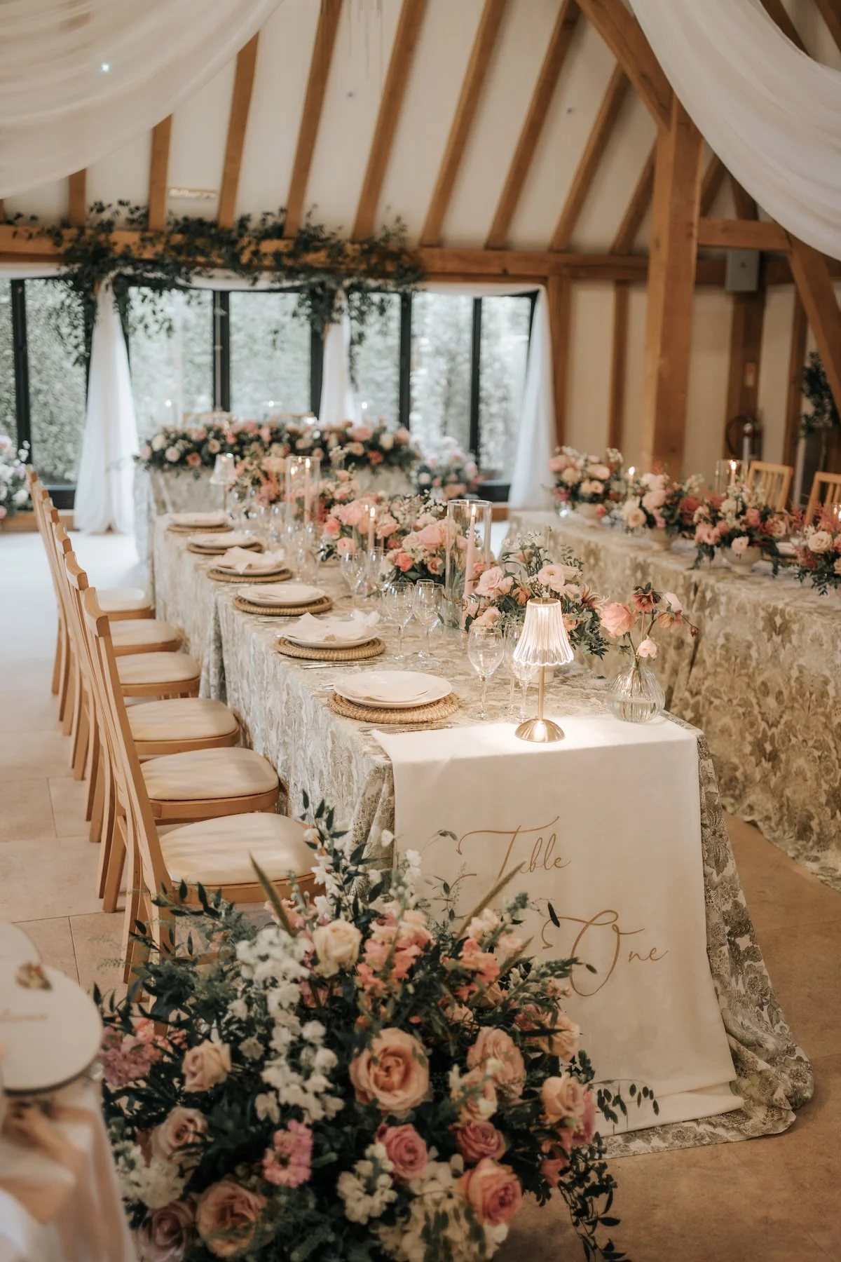 A long banquet table set for a wedding reception with floral arrangements, candles, and elegant tableware inside a wooden barn with large windows and draped white fabric.
