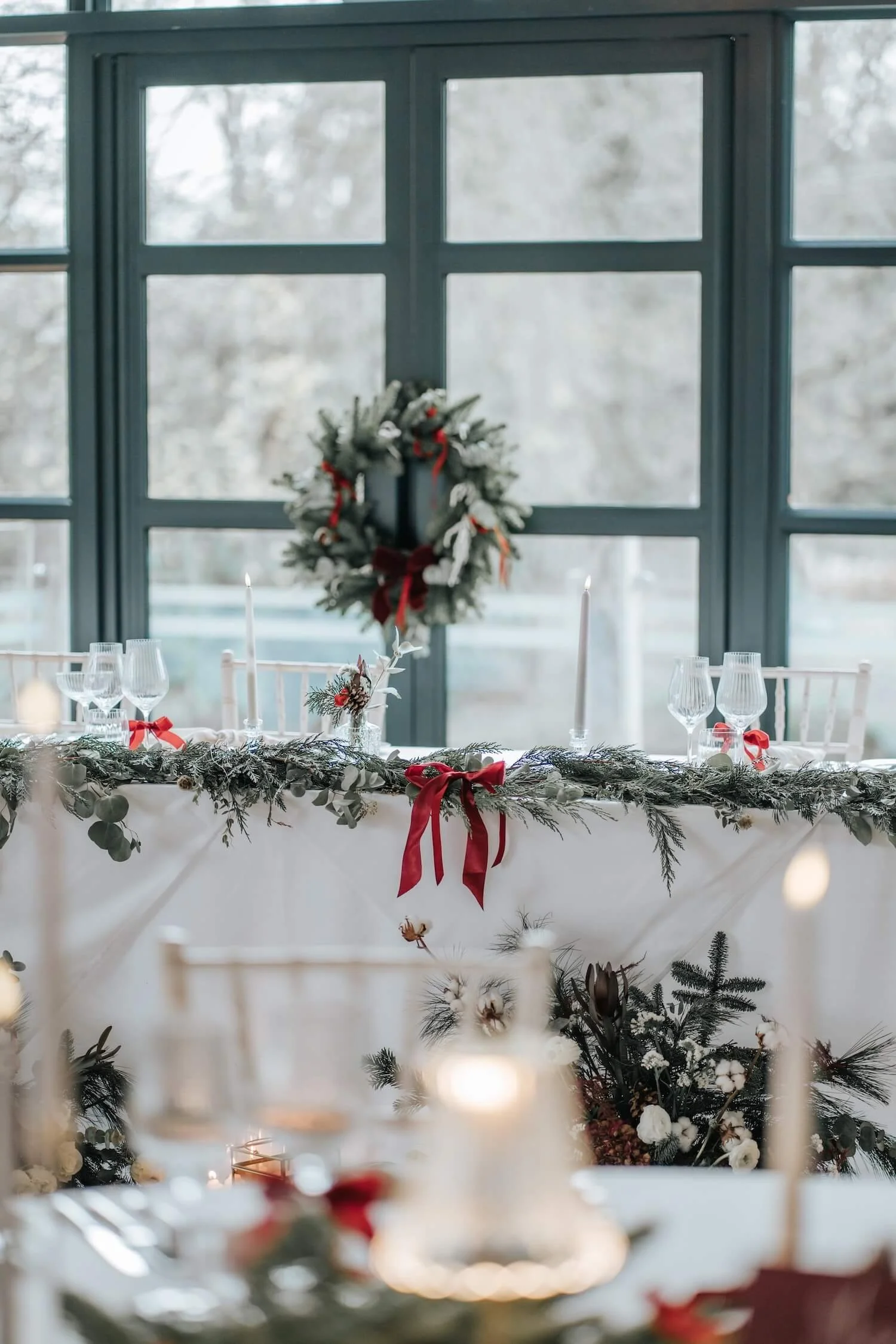 A decorated holiday table with a wreath on the window, candles, and greenery, set against a large window showing a wintery outdoor scene.