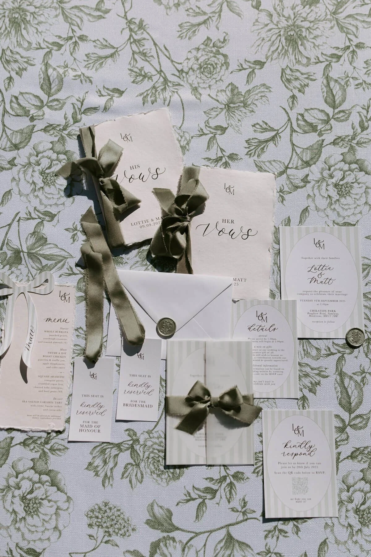 Wedding invitation suite on floral tablecloth. Includes invitations, RSVP card, menu, and additional notes, all with elegant script fonts and gray ribbons.