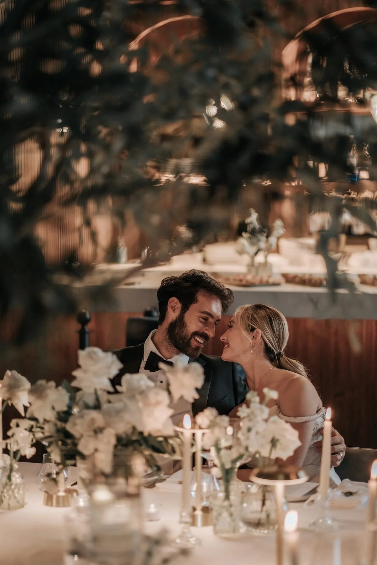 A couple at a wedding reception sitting at a decorated table with white flowers and candles, smiling and leaning close to each other.