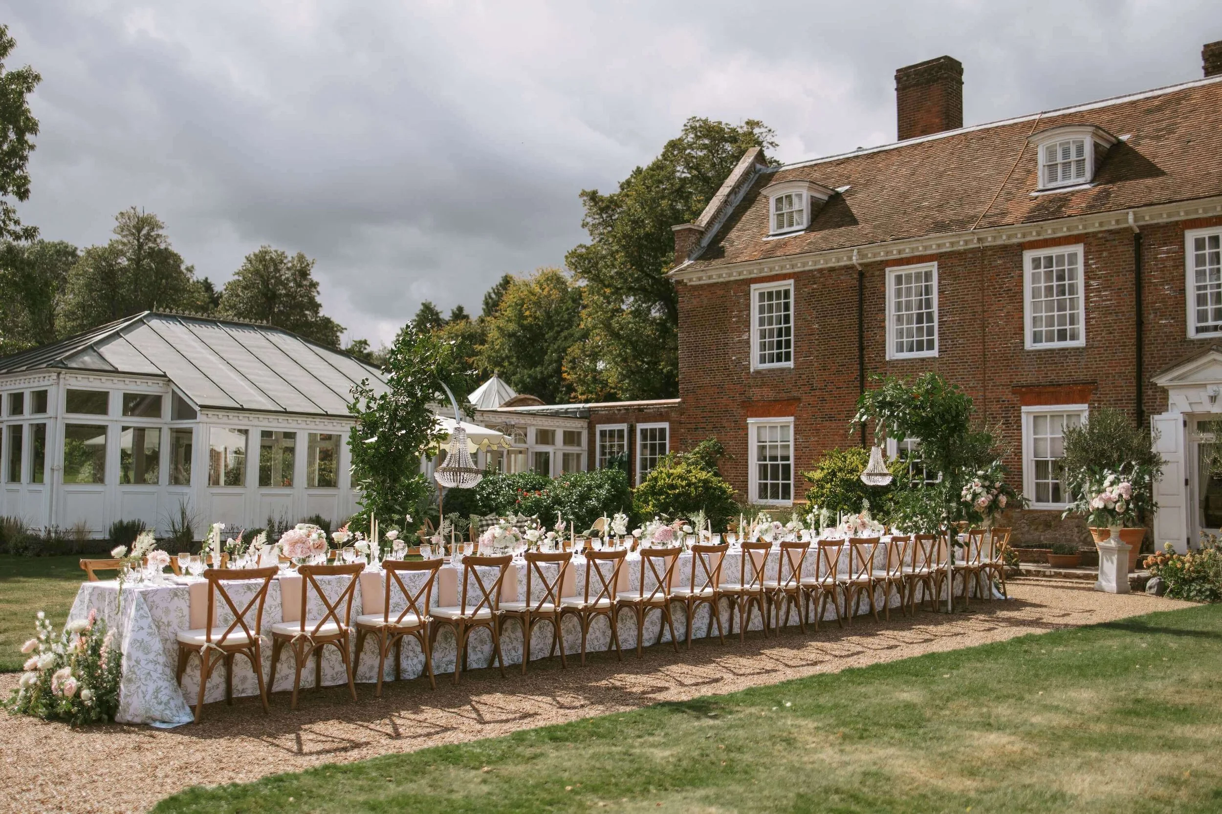 Outdoor wedding setup in front of a large brick house, with a long decorated table, chairs, floral arrangements, and a greenhouse structure nearby.