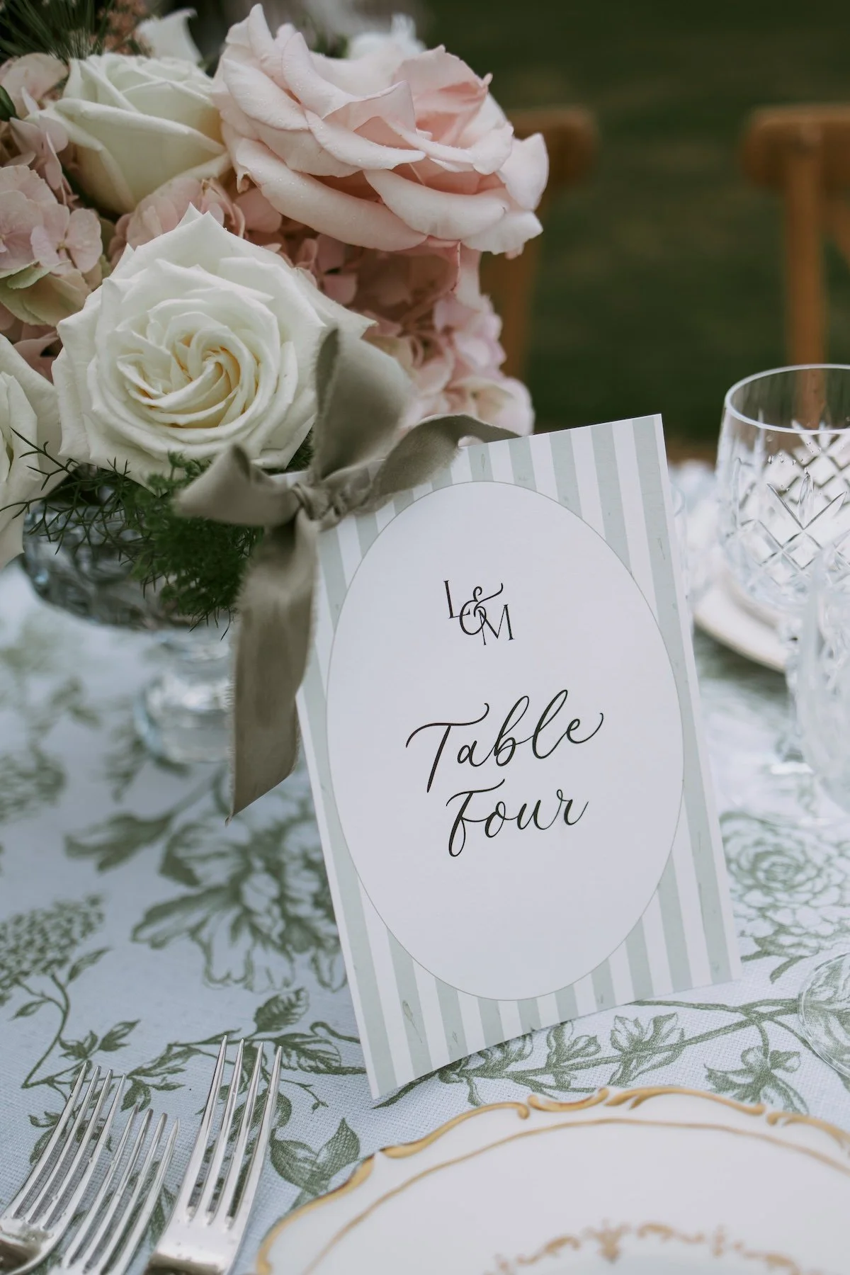 A wedding table setting with a floral centerpiece of white and light pink roses and hydrangeas, a table card labeled 'Table Four', crystal glassware, and a white and gold-rimmed plate, all arranged on a table with a patterned tablecloth.